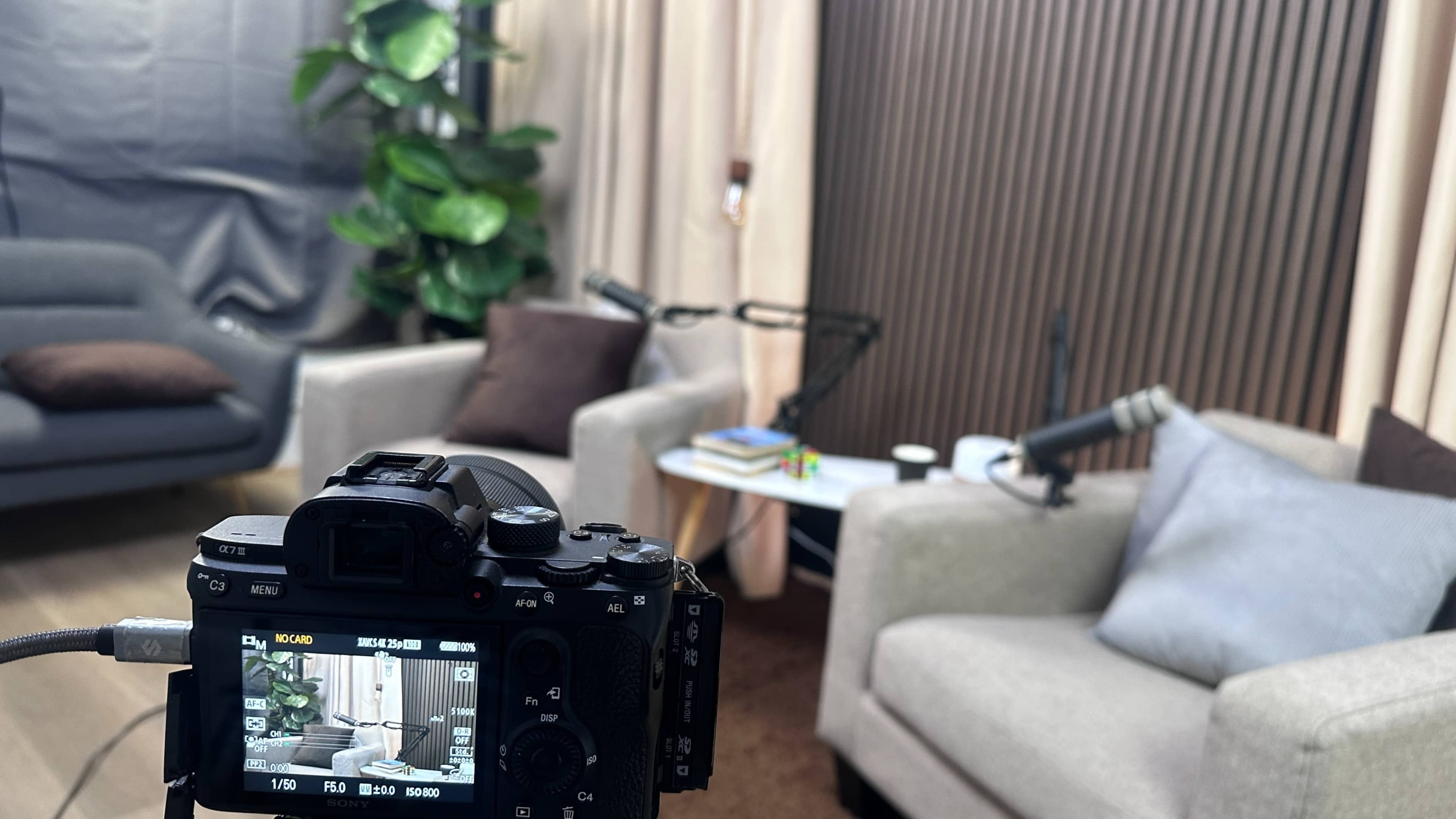 The image shows a camera set up in front of two chairs and a small table in a cozy studio with a plant and microphones.