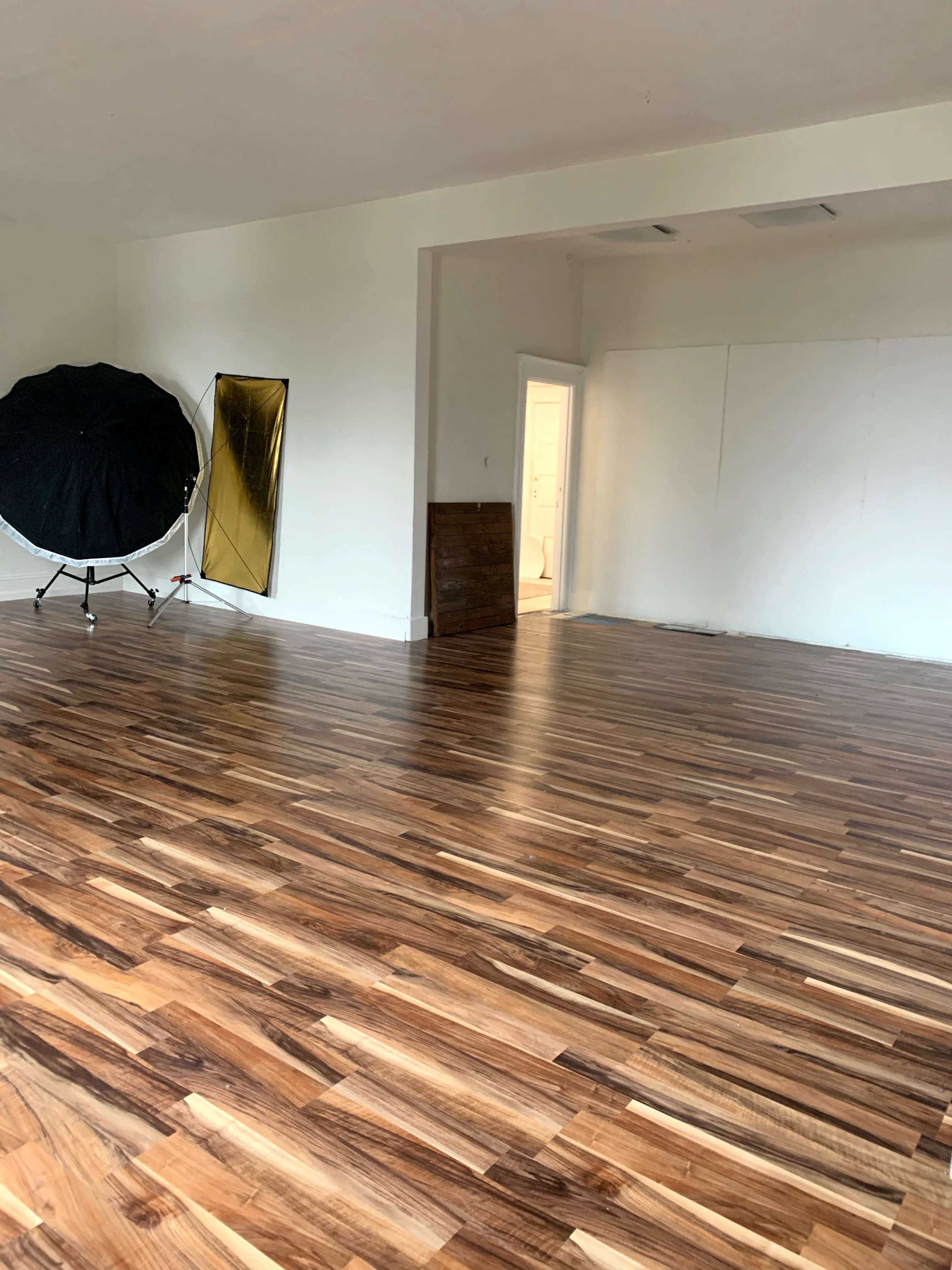 A spacious room with wooden flooring, a large reflector umbrella, a gold mirror on the wall, and an open doorway leading to another area.