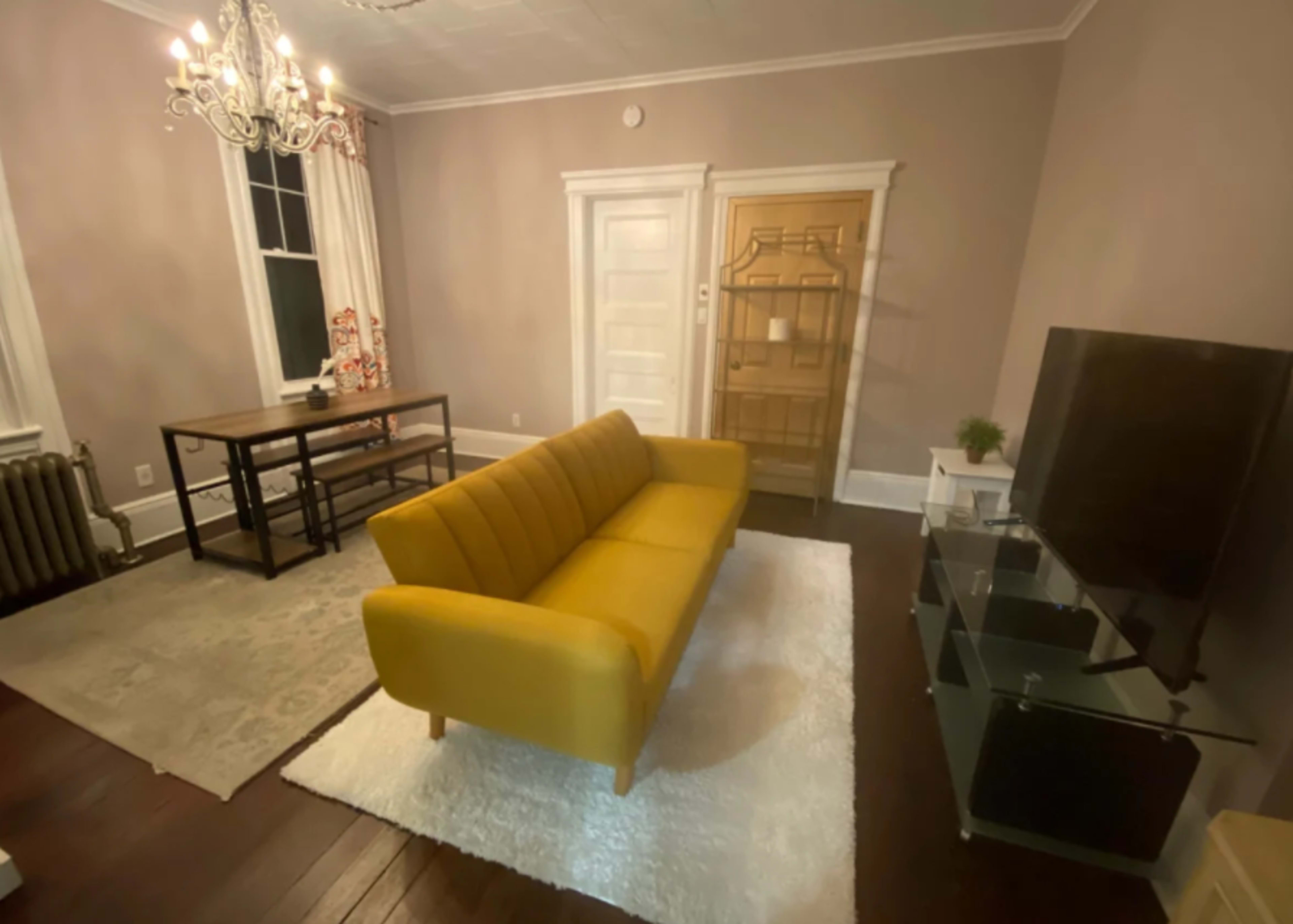 A yellow couch is positioned in a living room with a chandelier, a wooden table, a TV stand, and a door leading outside.