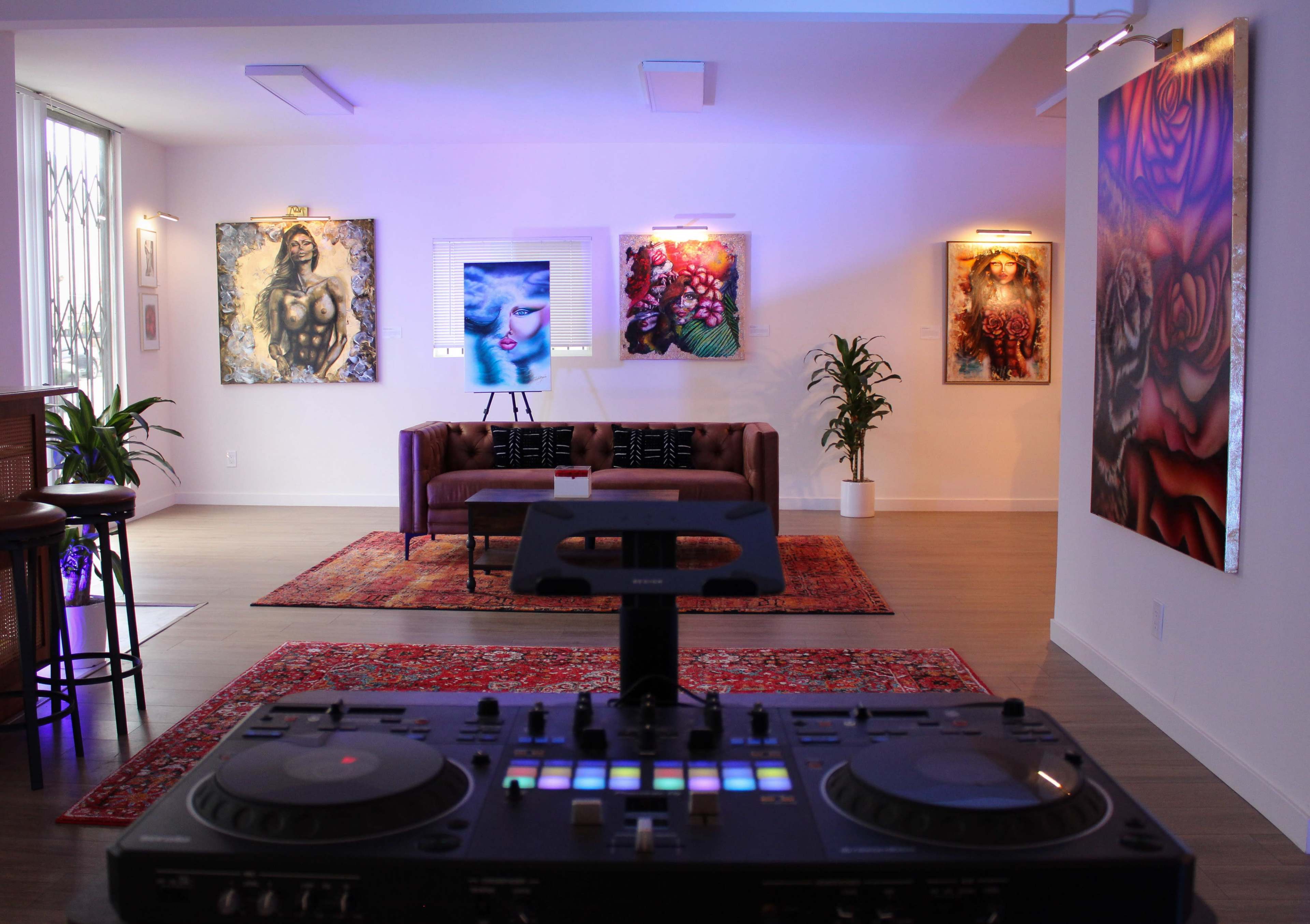 The image shows a modern art gallery featuring vibrant paintings on the walls, a brown couch in the center, and a DJ turntable in the foreground.