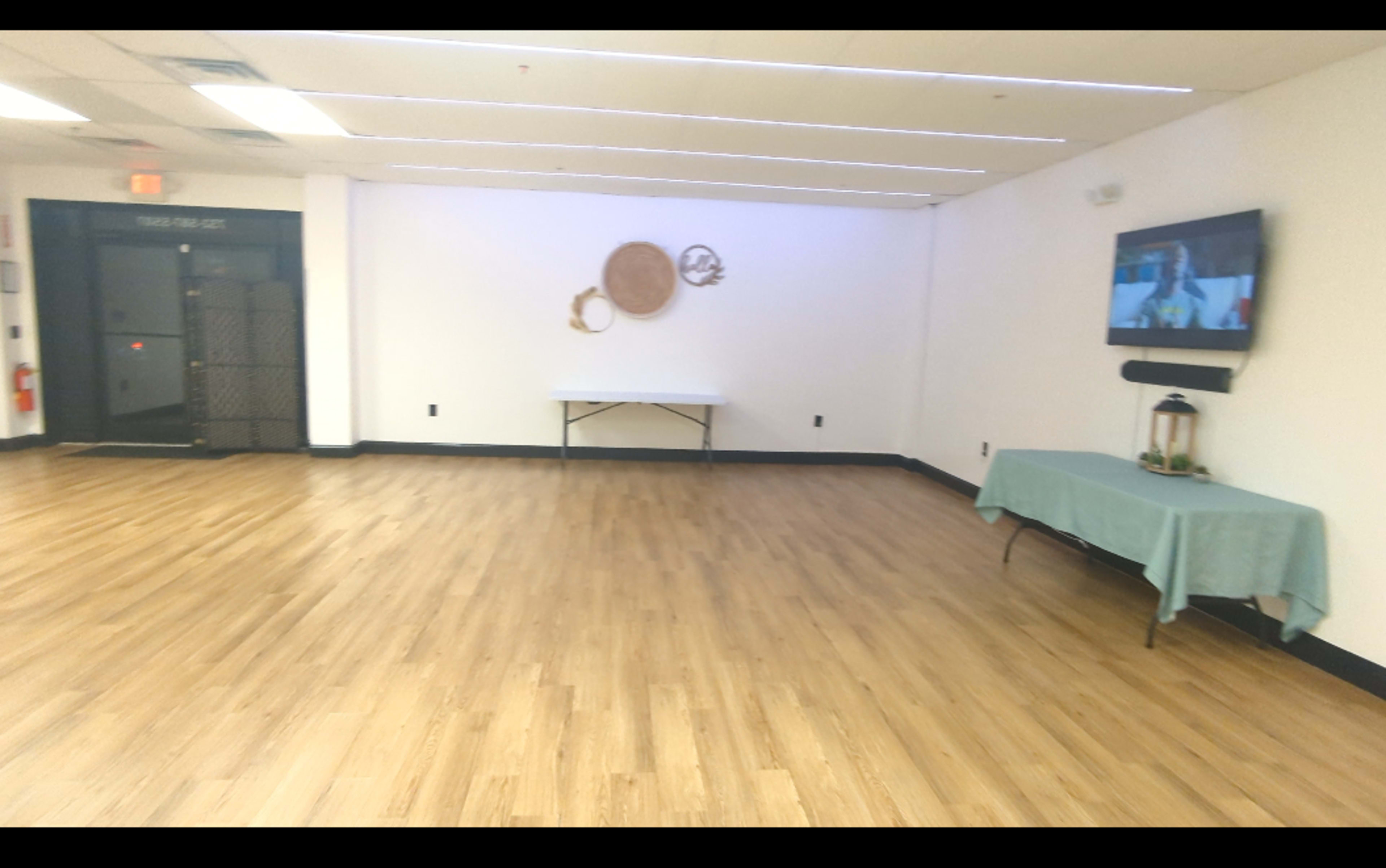 The image shows a spacious, empty room with wooden flooring, featuring a table covered with a light blue cloth and a wall-mounted television.