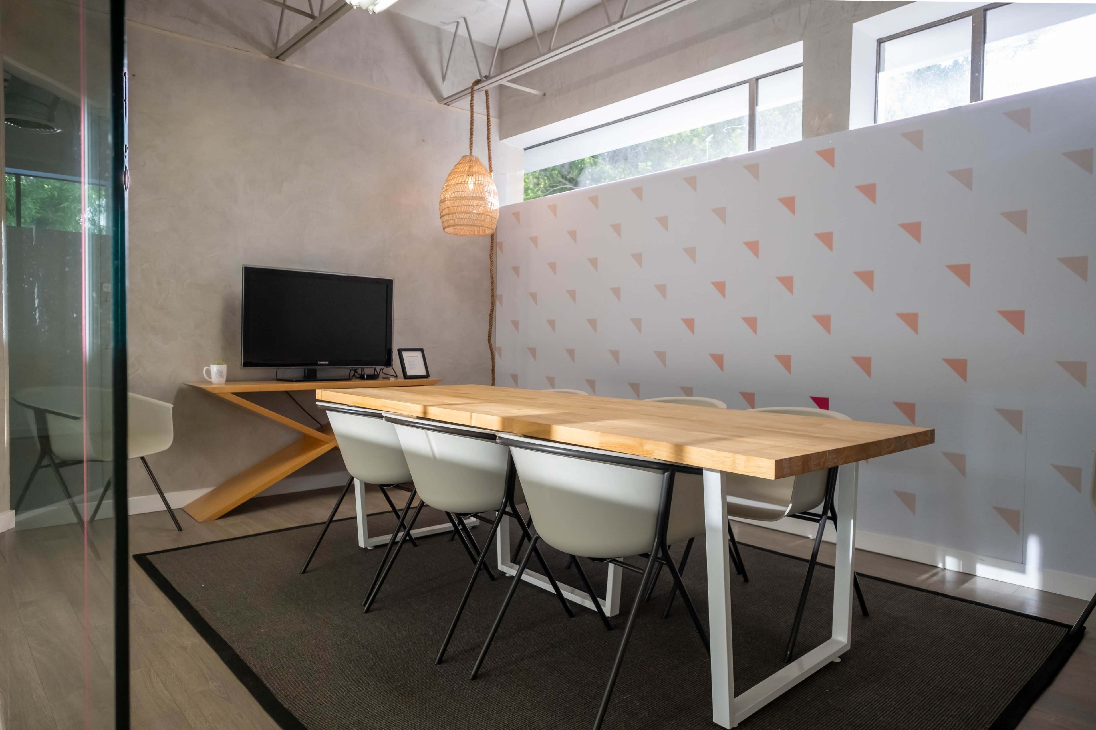 The image shows a modern meeting room with a wooden table, six white chairs, a television on a stand, and a decorative wall featuring a geometric design.
