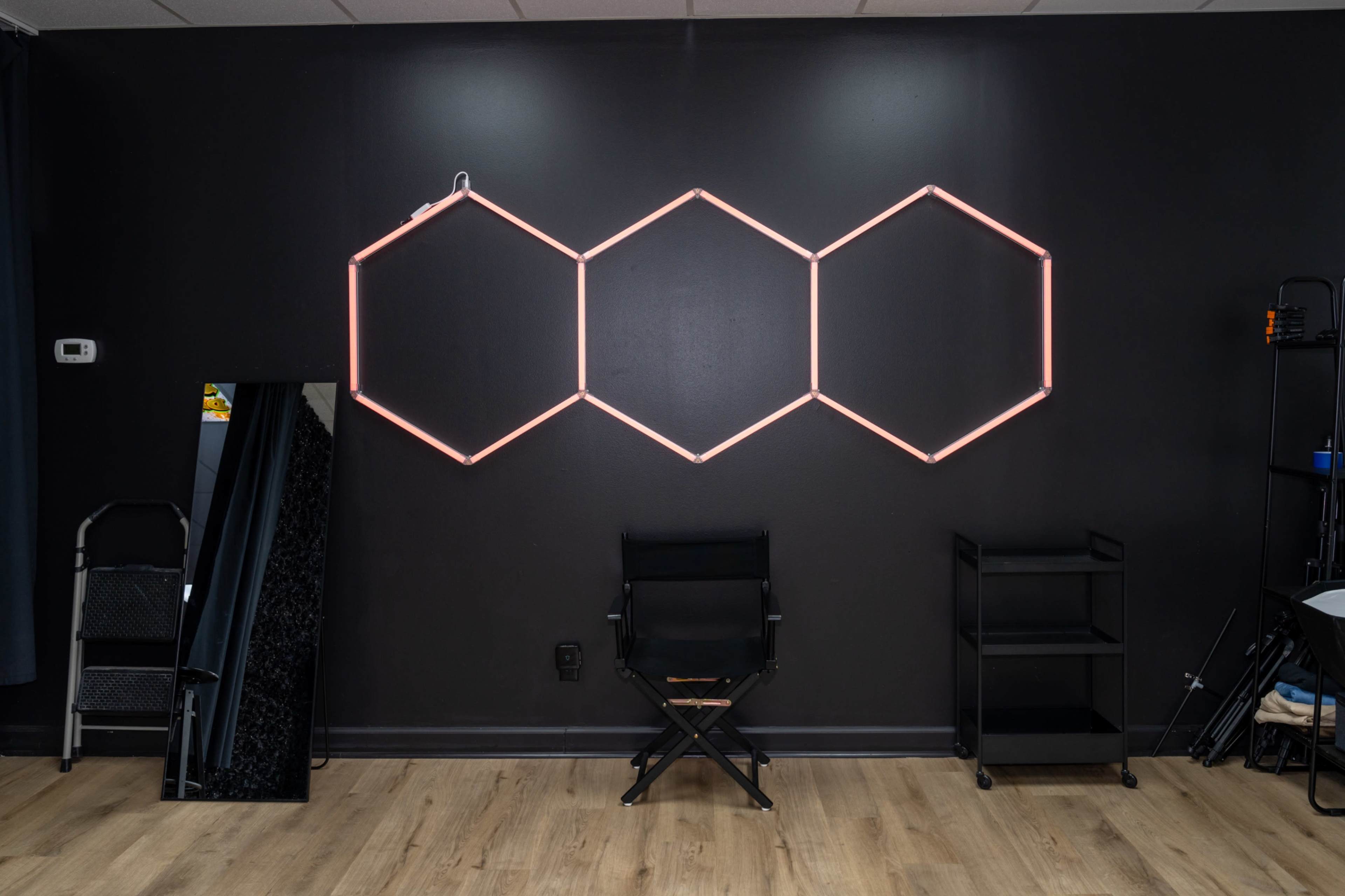 The image shows a black wall with three hexagonal neon light fixtures arranged above a chair and a mirror.