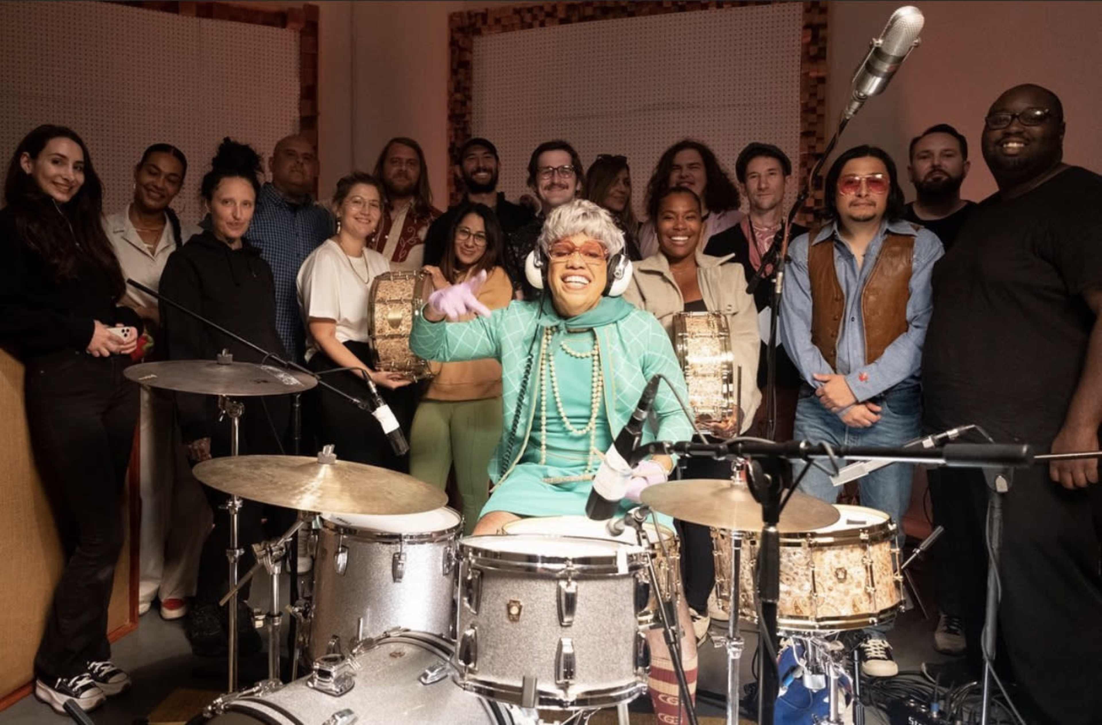 A group of musicians poses together in a recording studio, with a drummer in front smiling and playing the drums.