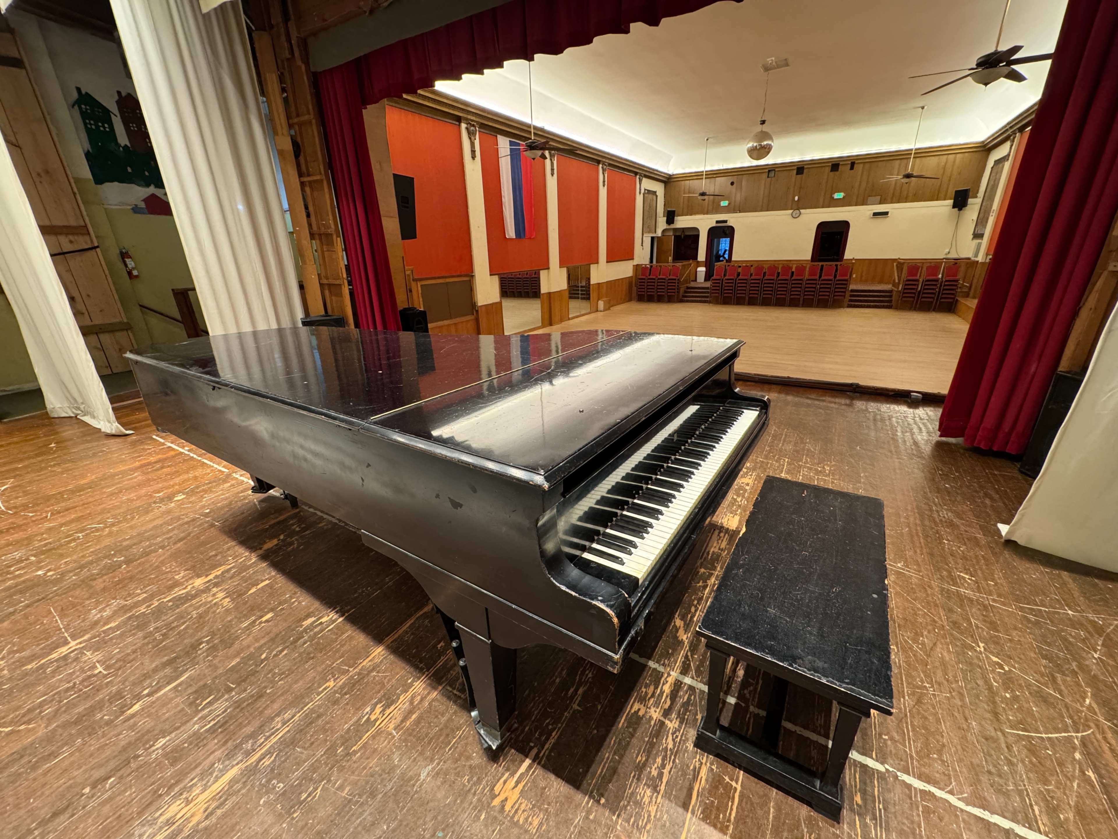 A black grand piano sits next to a small bench in an empty auditorium with wooden flooring and red curtains.