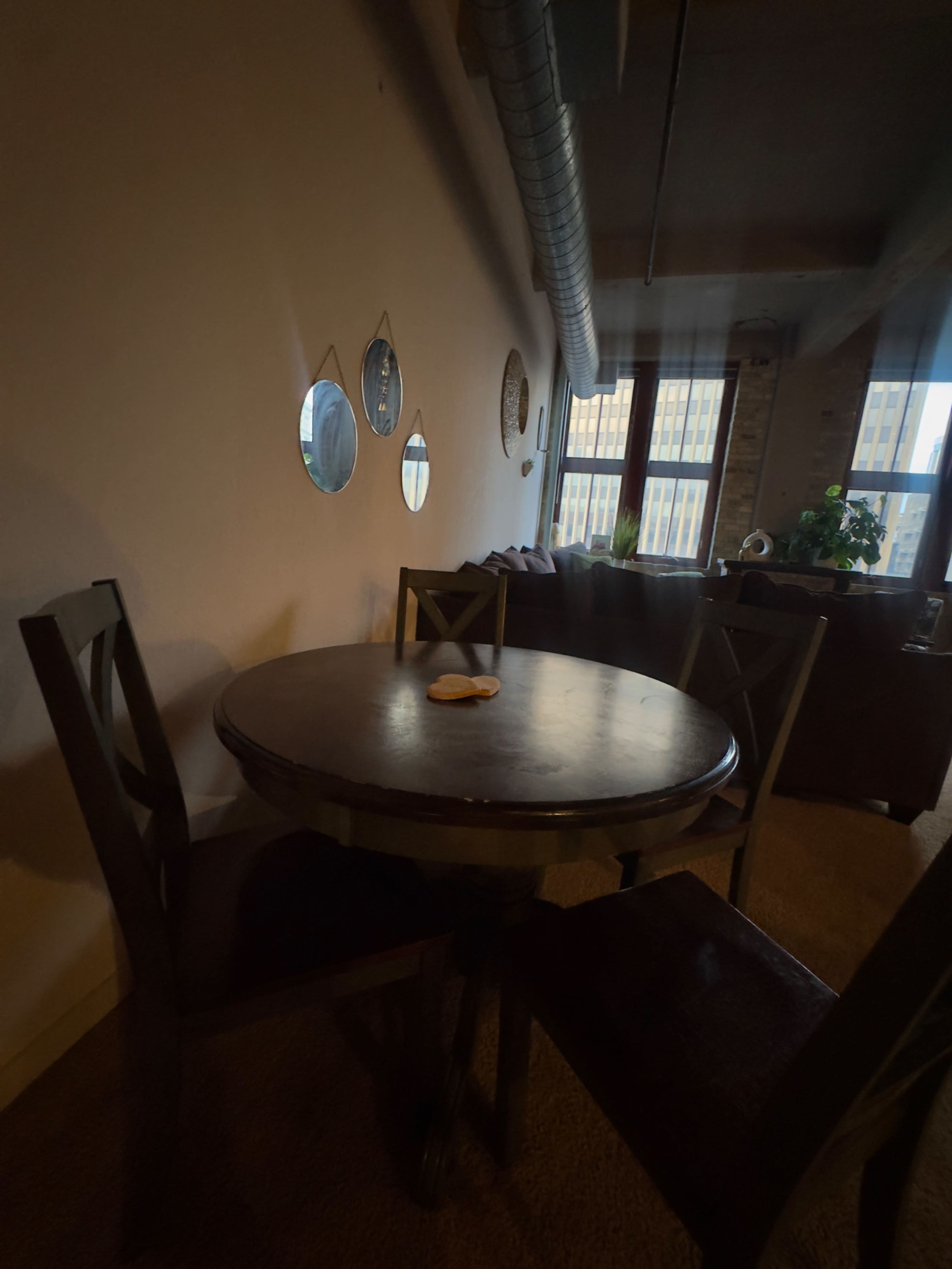 A round wooden table with two chairs sits in a well-lit room, featuring small round mirrors on the wall and a view of a cityscape through large windows.