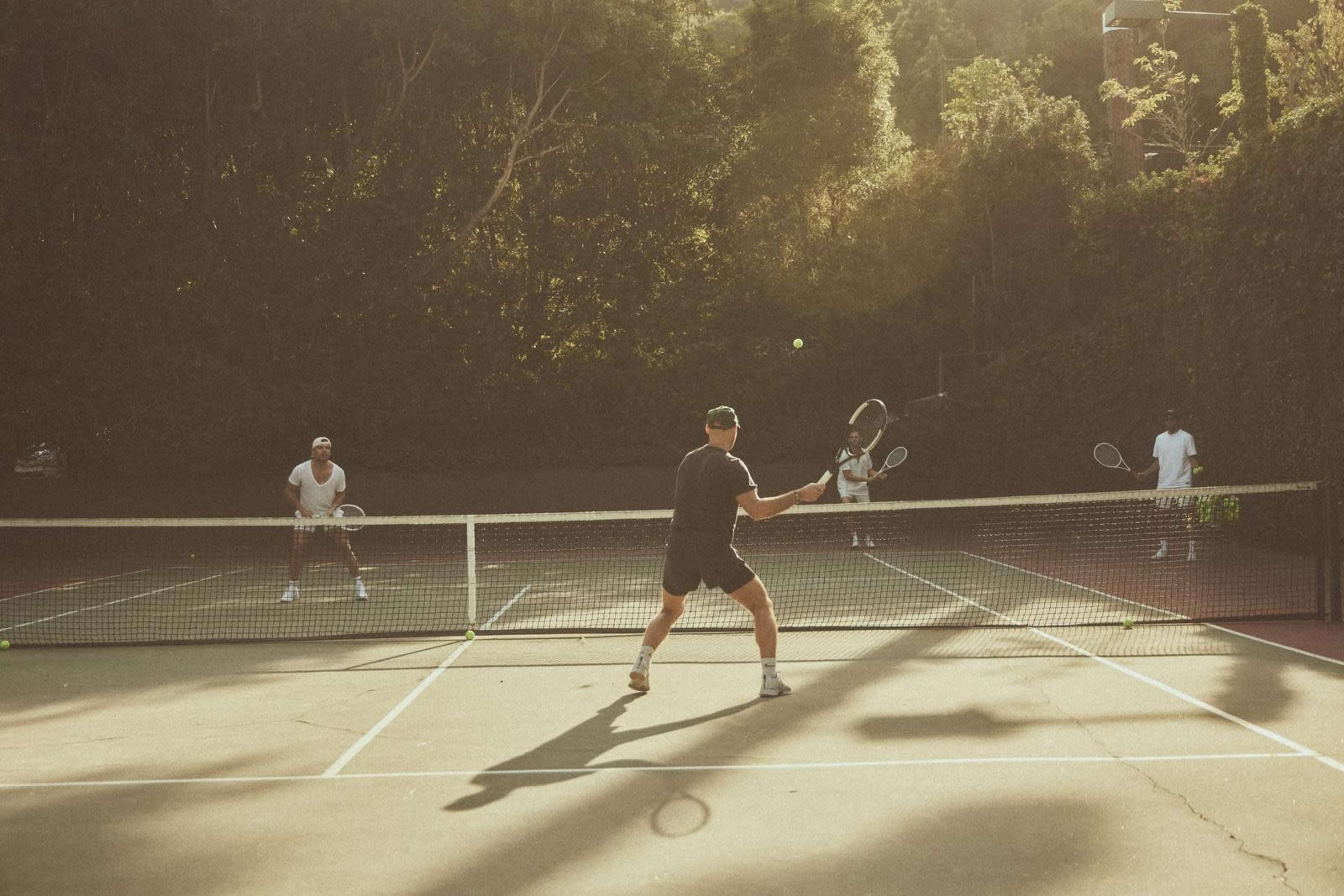 A group of three men plays tennis on an outdoor court surrounded by greenery.