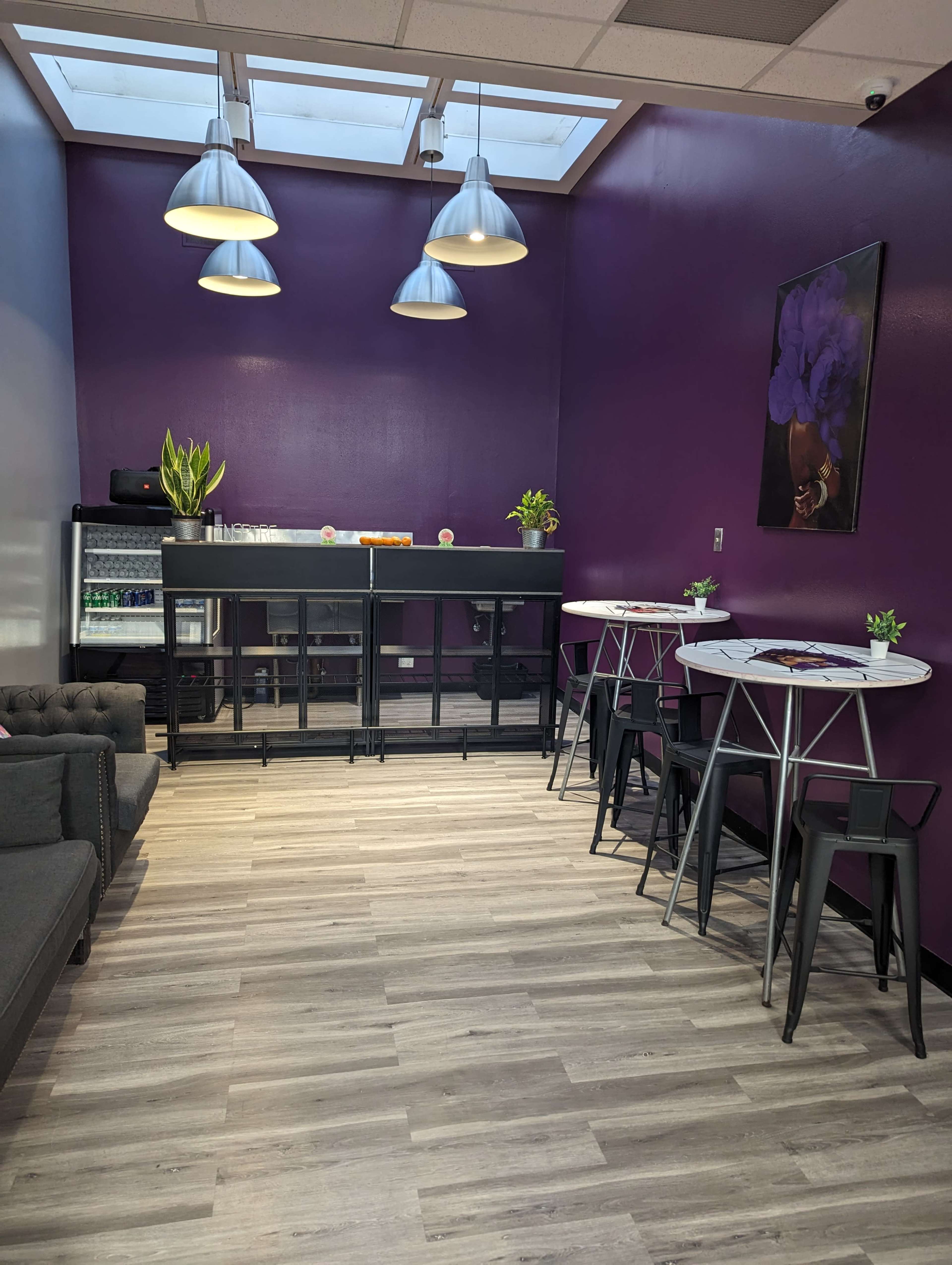 A small, modern seating area with purple walls, a few tall tables and chairs, a gray couch, and plants, illuminated by pendant lights.