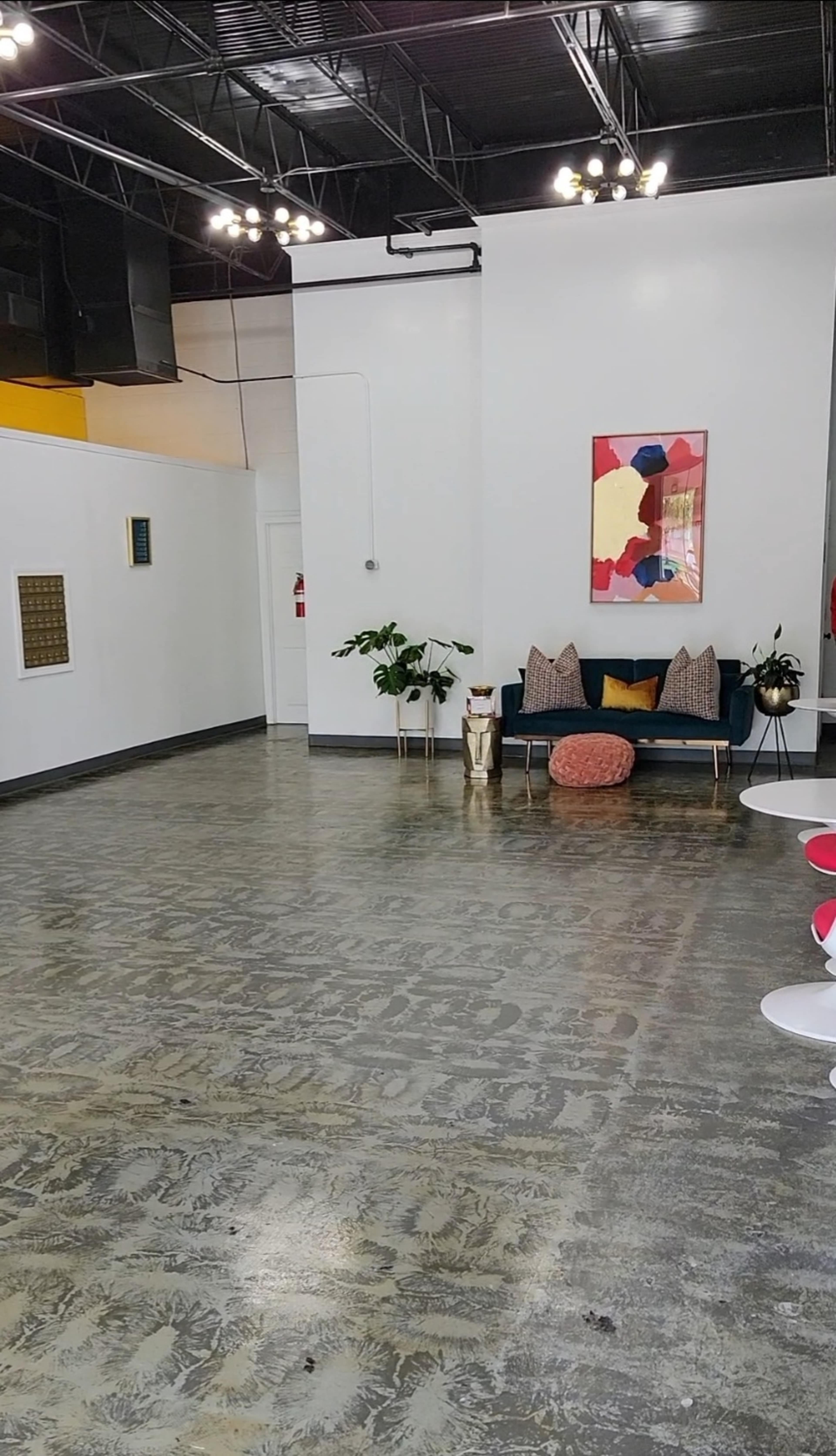 A spacious, well-lit interior with a sofa, decorative pillows, a plant, and a round orange pouf, alongside a coffee table and artwork on the wall.