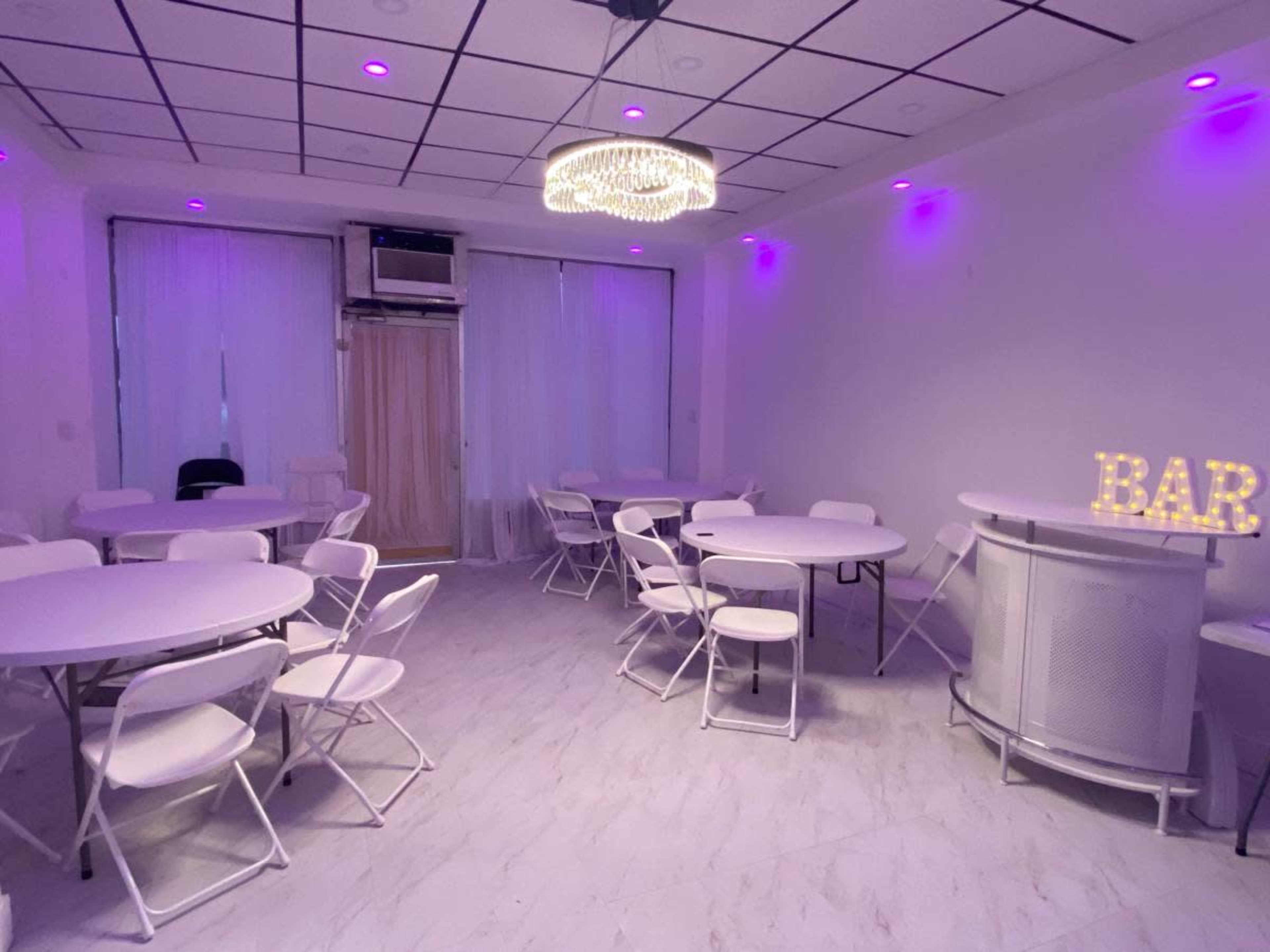 A spacious room features white folding tables and chairs, a bar counter, and purple LED lighting.