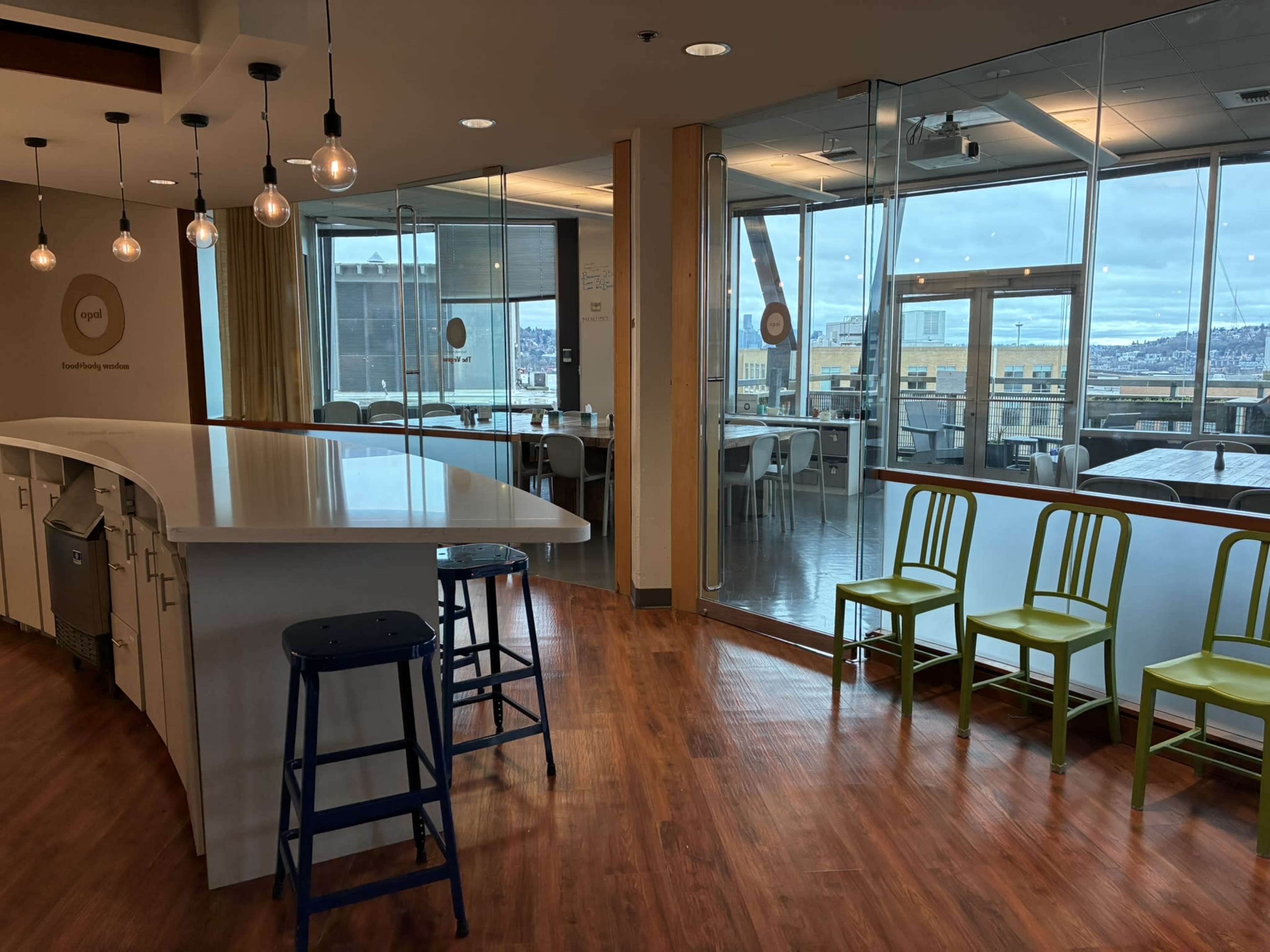 The image shows a modern interior space with a bar-style countertop, a few stools, and bright green chairs, all set against large windows overlooking a cityscape.