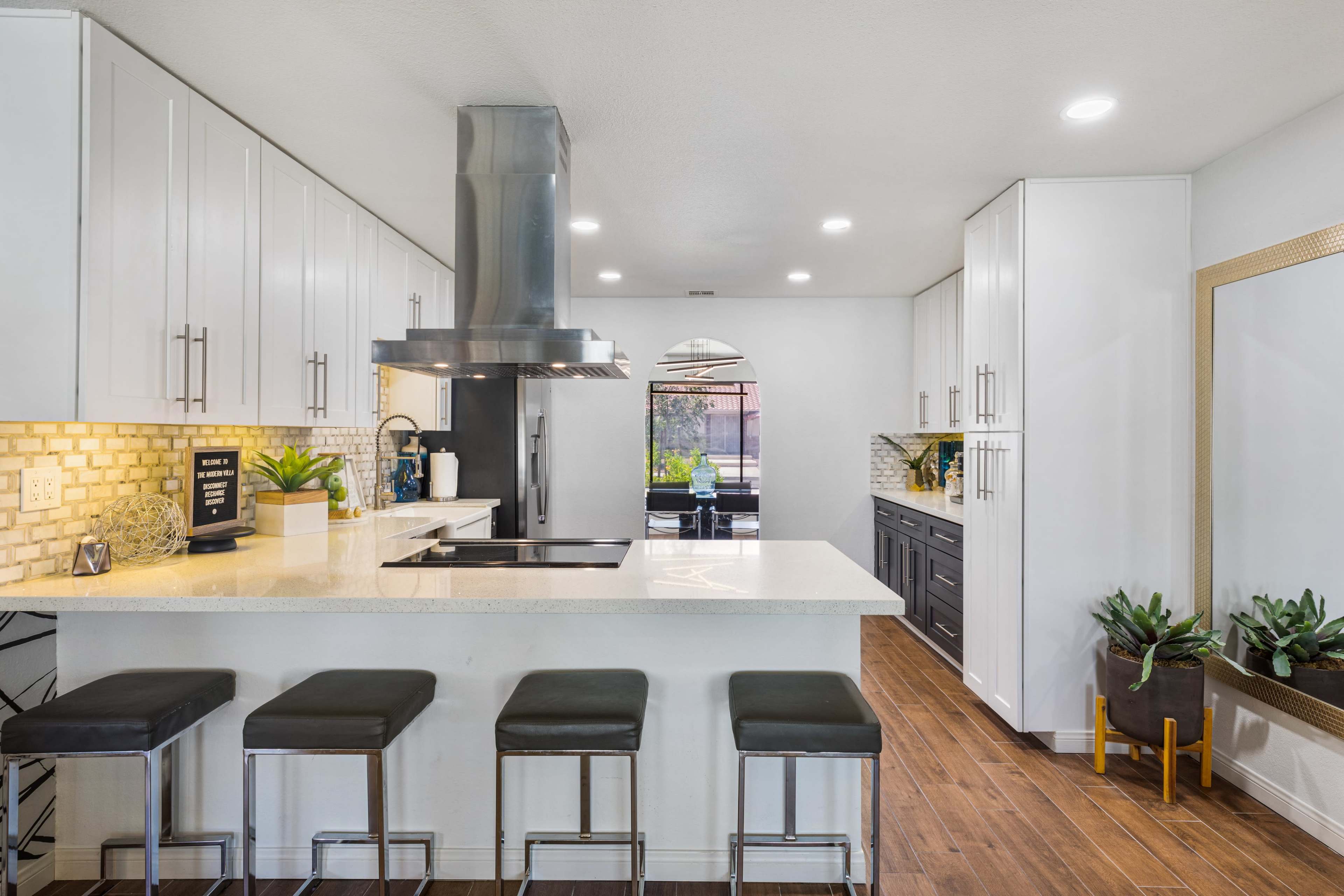 A modern kitchen features a central island with bar stools, stainless steel appliances, and dark cabinetry against light walls.