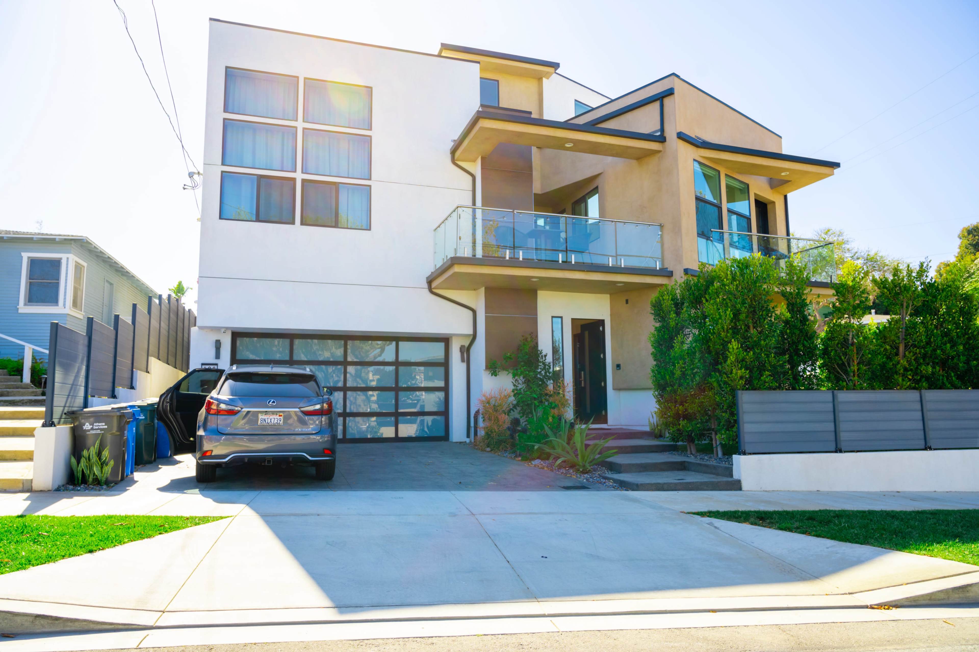 A modern two-story house with large windows, a balcony, a landscaped front yard, and a car parked in the driveway.