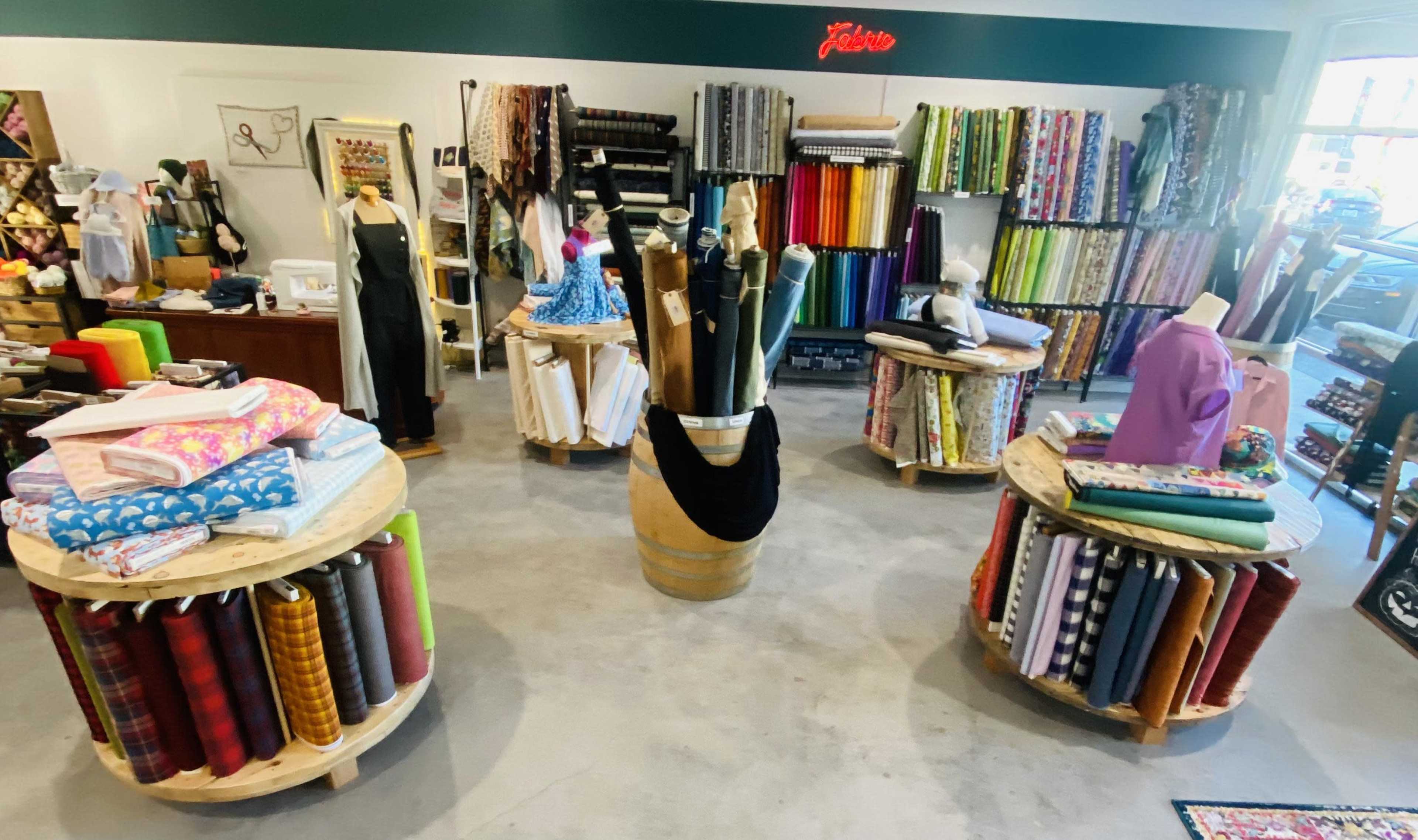 The image shows a fabric store with various colorful rolls of fabric displayed on circular wooden tables and shelves lining the walls.