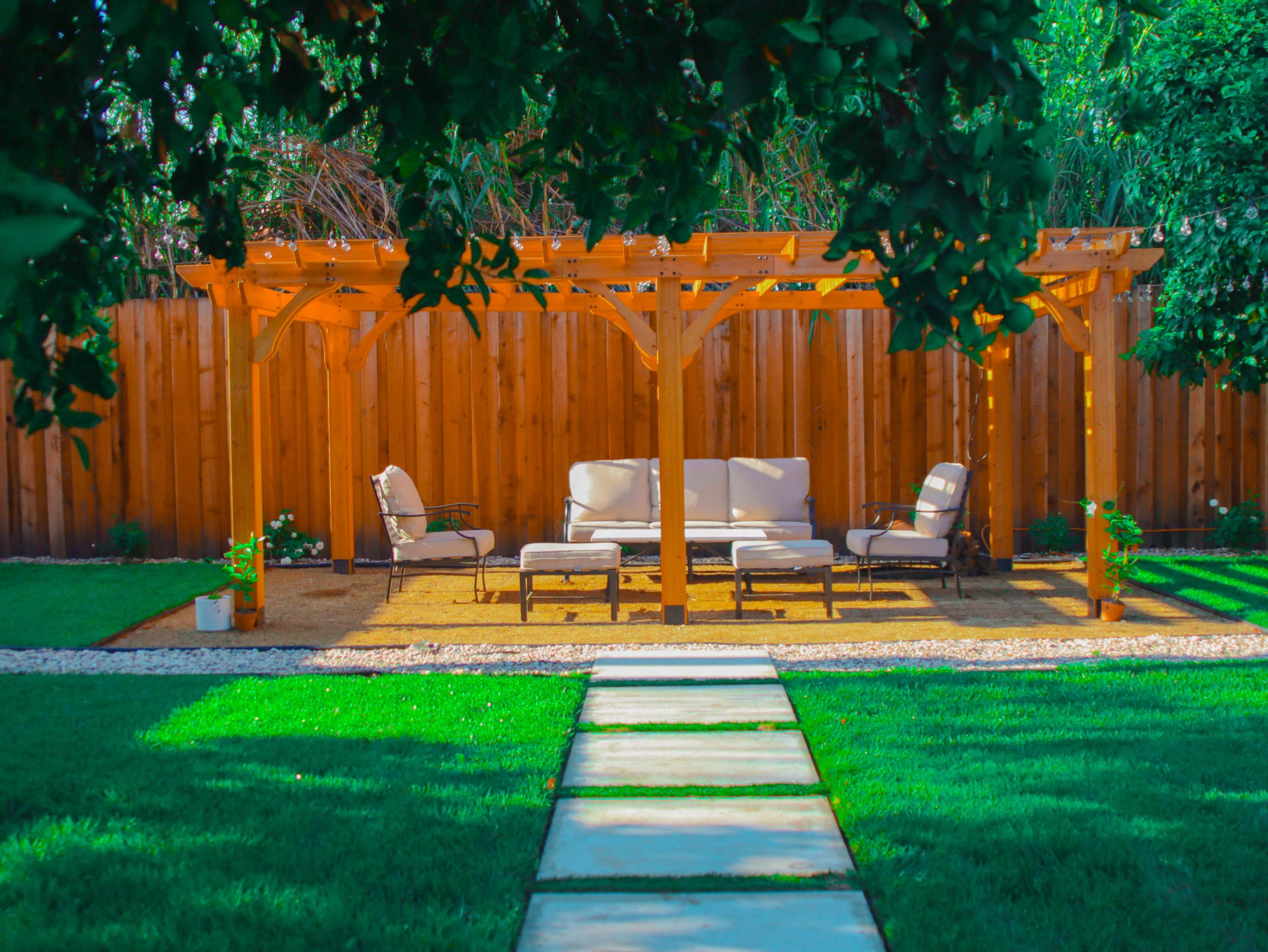 A wooden pergola provides shade over a set of beige outdoor furniture in a landscaped backyard featuring a stone pathway and well-manicured grass.