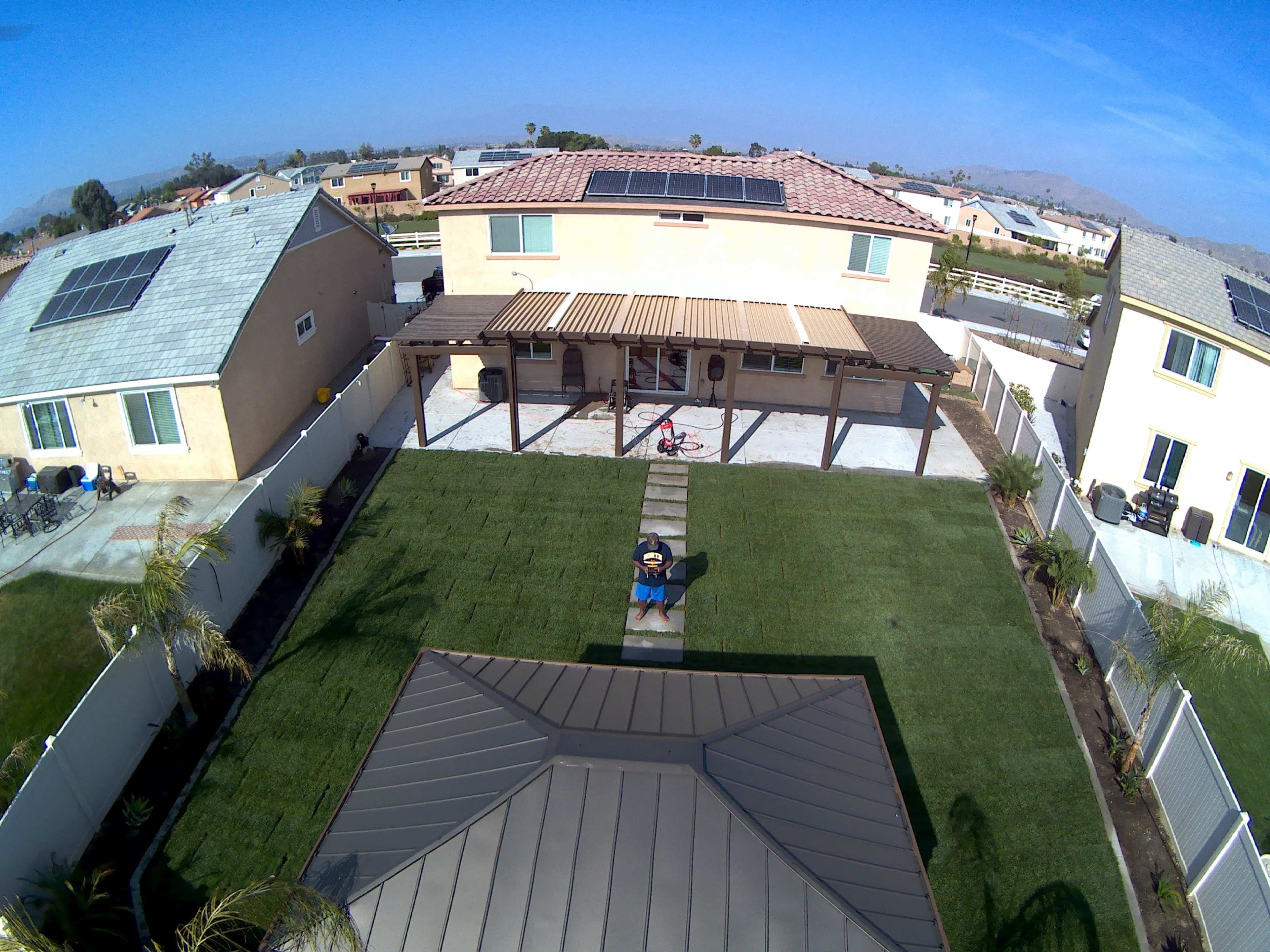 The image shows a neighborhood with several houses, a green lawn, pathways, and solar panels on the roofs.