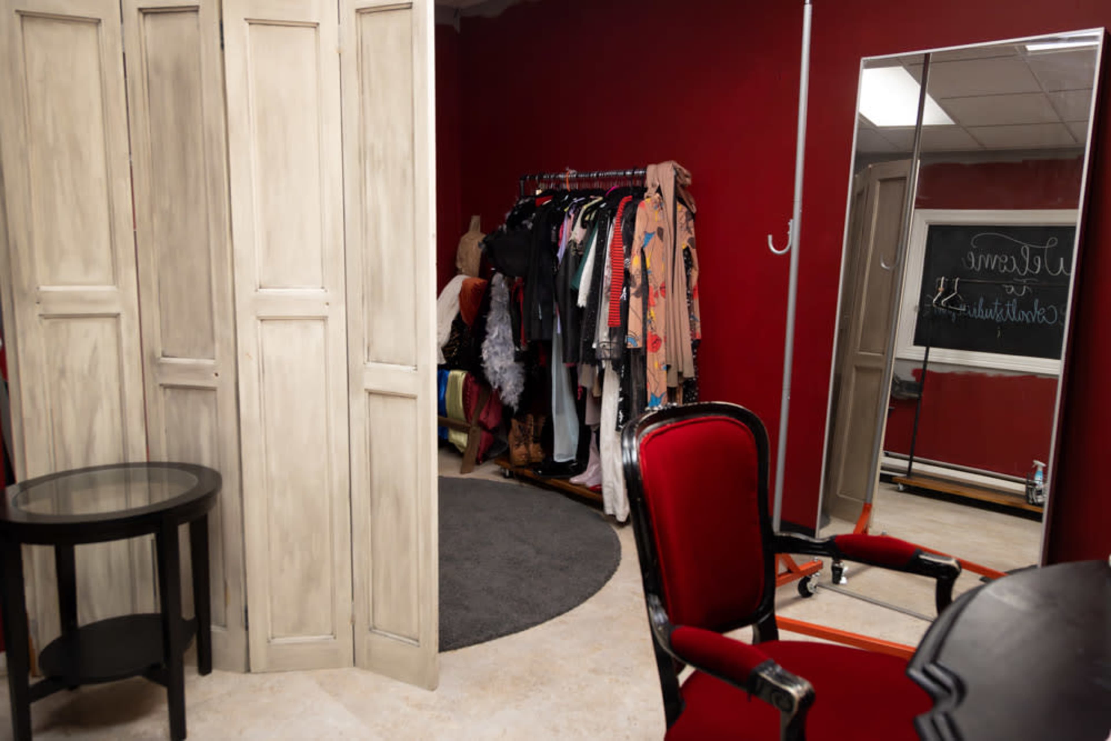 The image shows a dressing room with a red wall, a clothing rack filled with various garments, a full-length mirror, and a red upholstered chair beside a small round table.