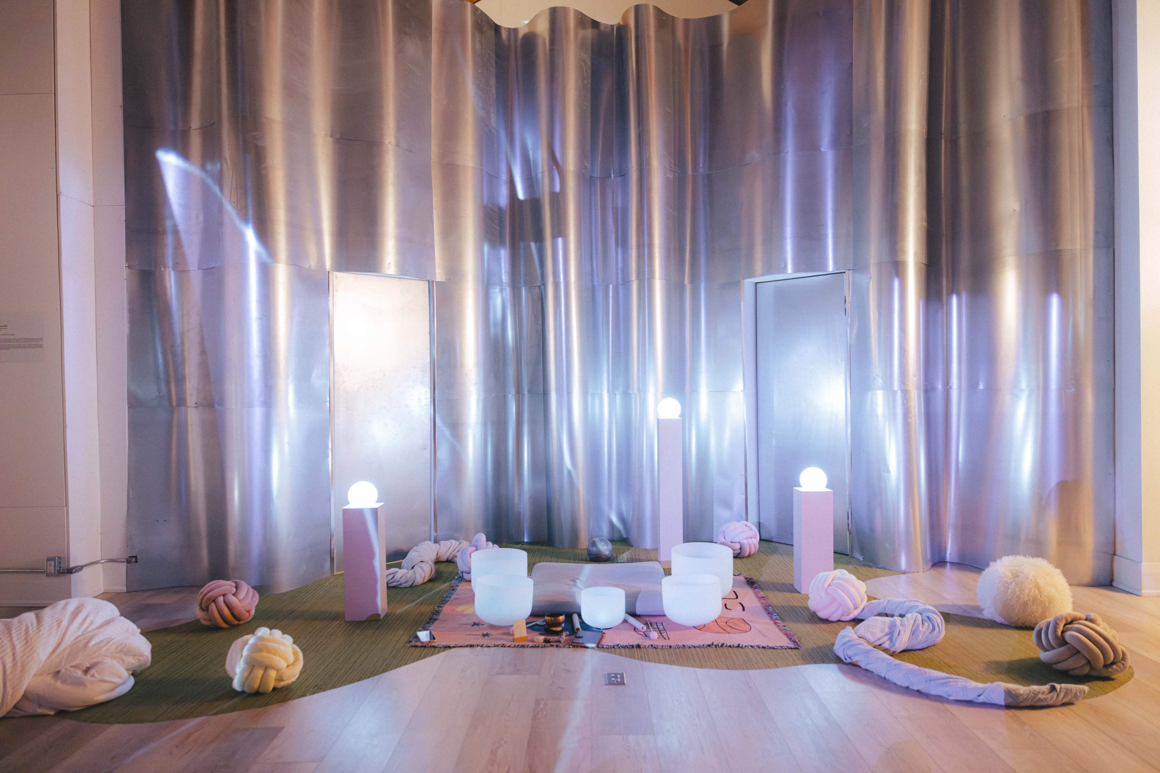 The scene features a contemporary art installation with metallic walls, glowing orbs, and various soft cushions scattered on the floor.