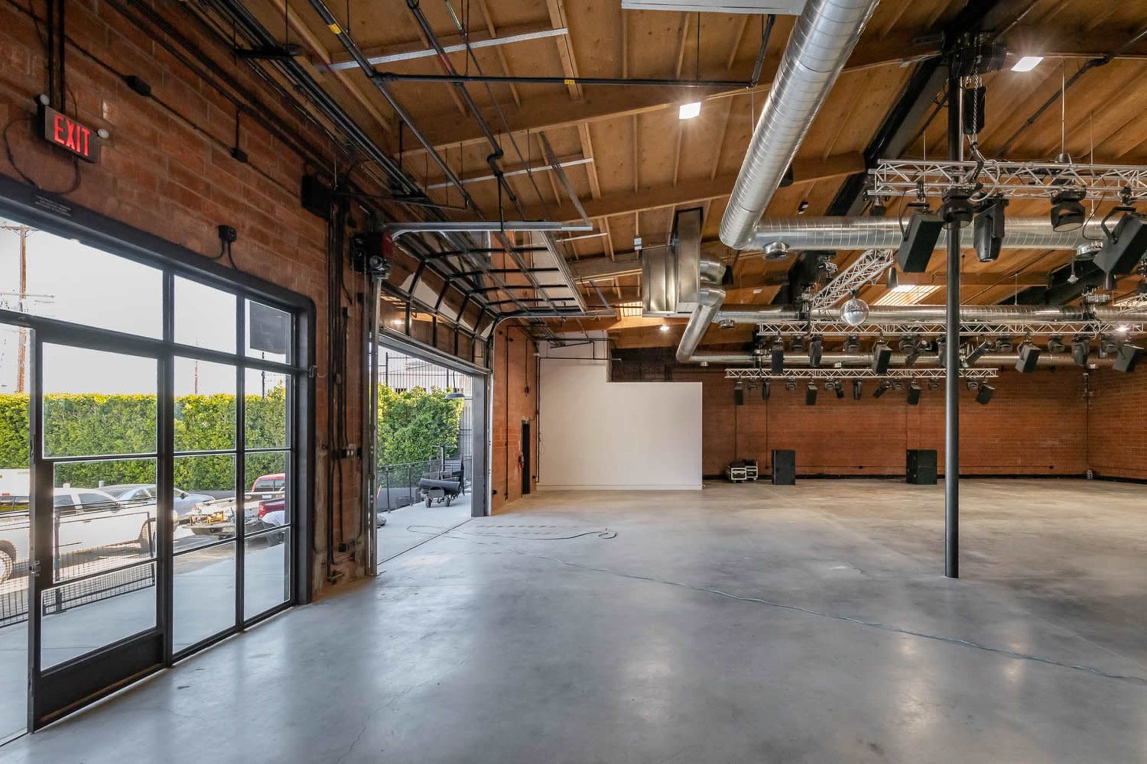 An empty industrial-style event space with exposed brick walls, wooden beams, and large windows leading to an outdoor area.
