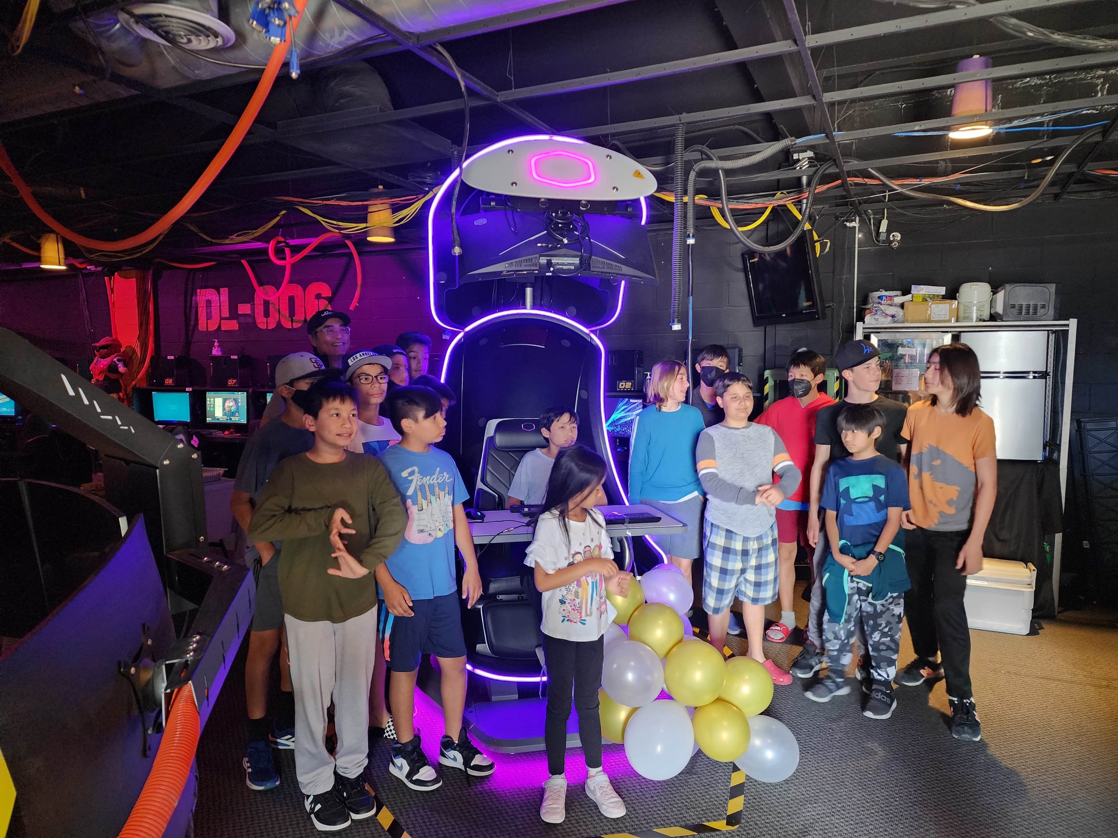A group of children and adults stands around a brightly lit gaming chair in a dark room filled with computer stations.