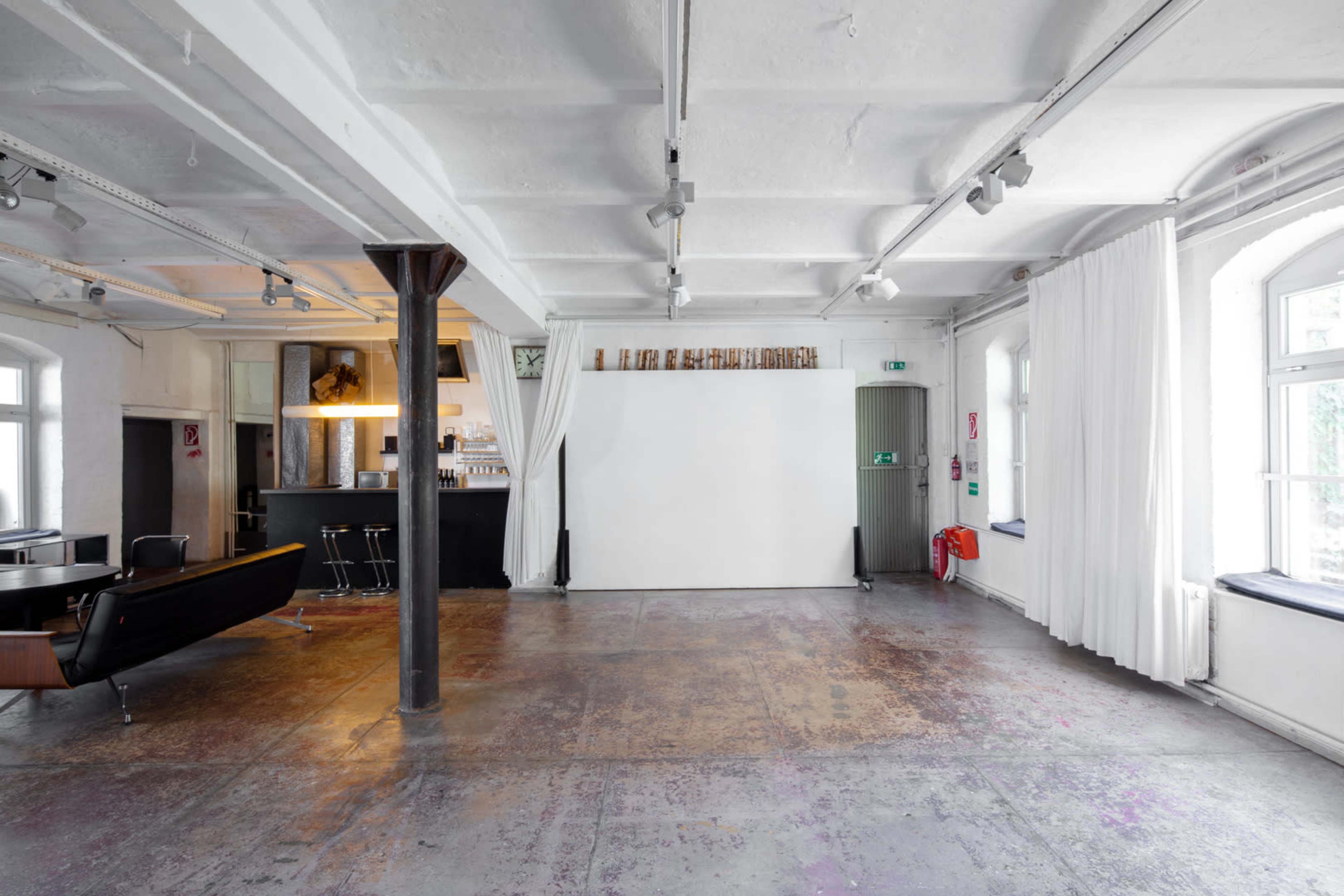 The image shows a spacious, minimalistic interior with a concrete floor, large windows, and a blank wall, accompanied by elements like a bar and seating area.