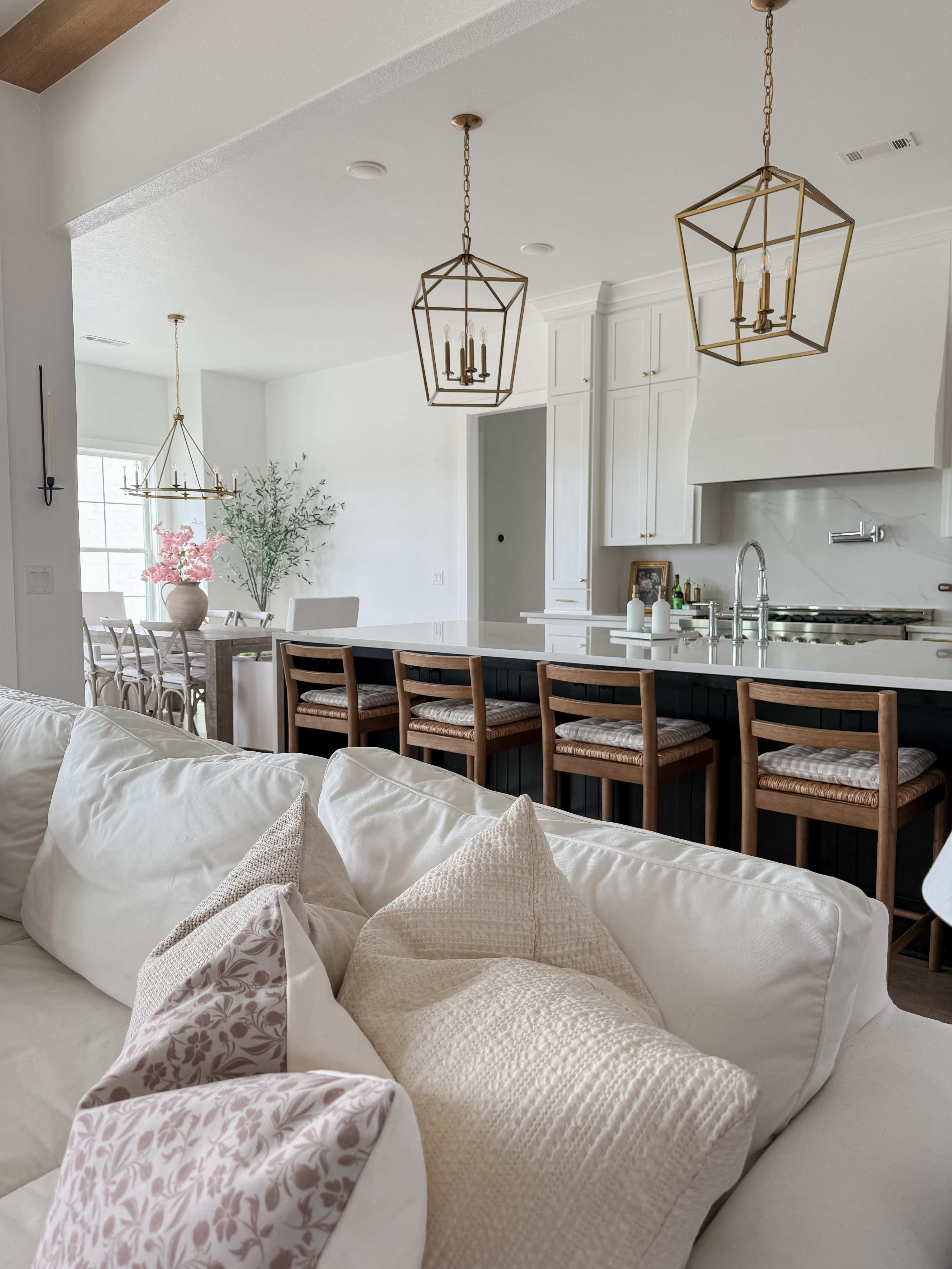 The image shows a modern kitchen and dining area with a white couch in the foreground, and three pendant lights hanging above a dark island bar with wooden stools.