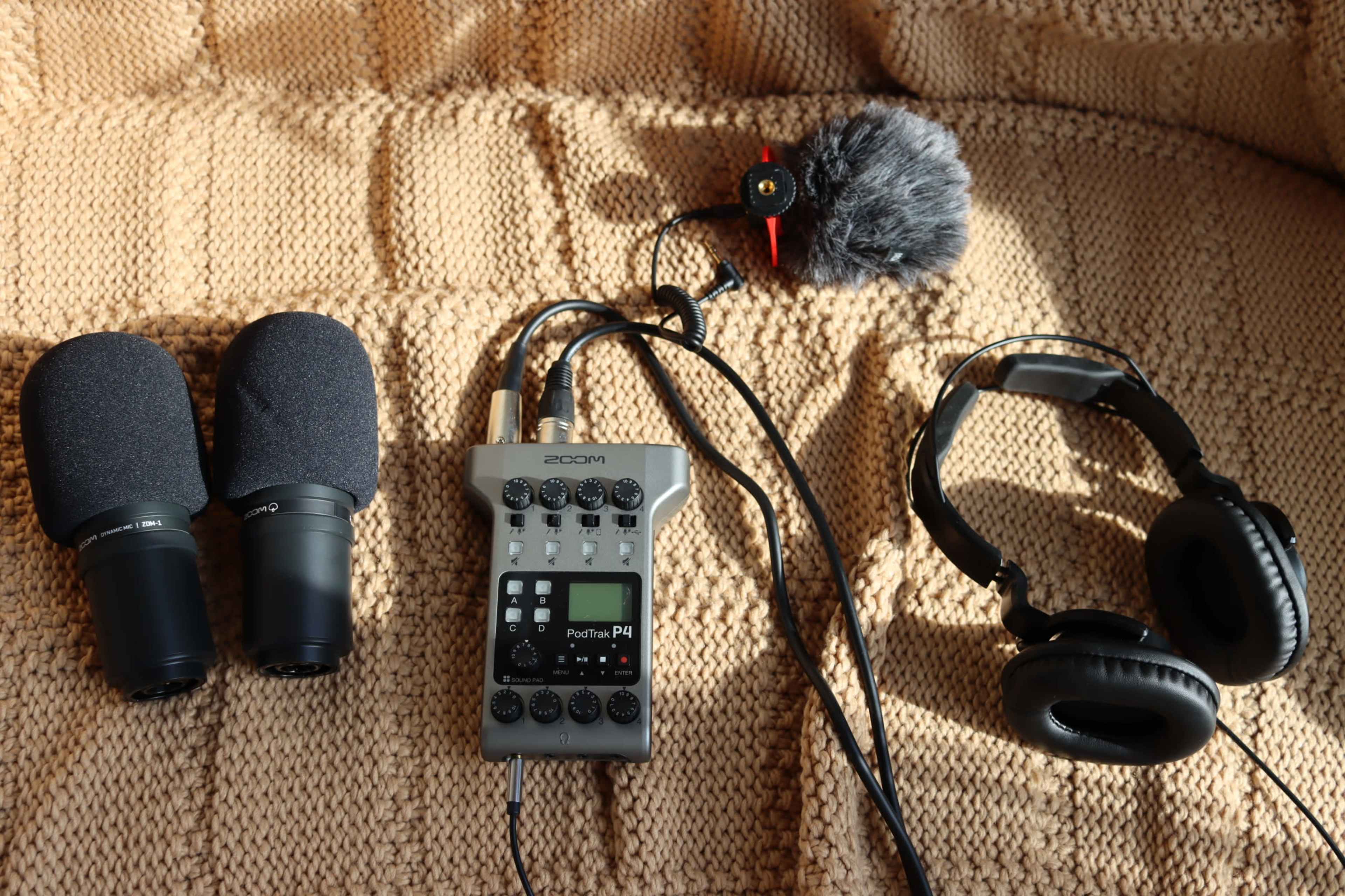 Two microphones, a portable audio recorder, a fuzzy microphone windscreen, and a pair of headphones arranged on a textured, beige fabric surface.