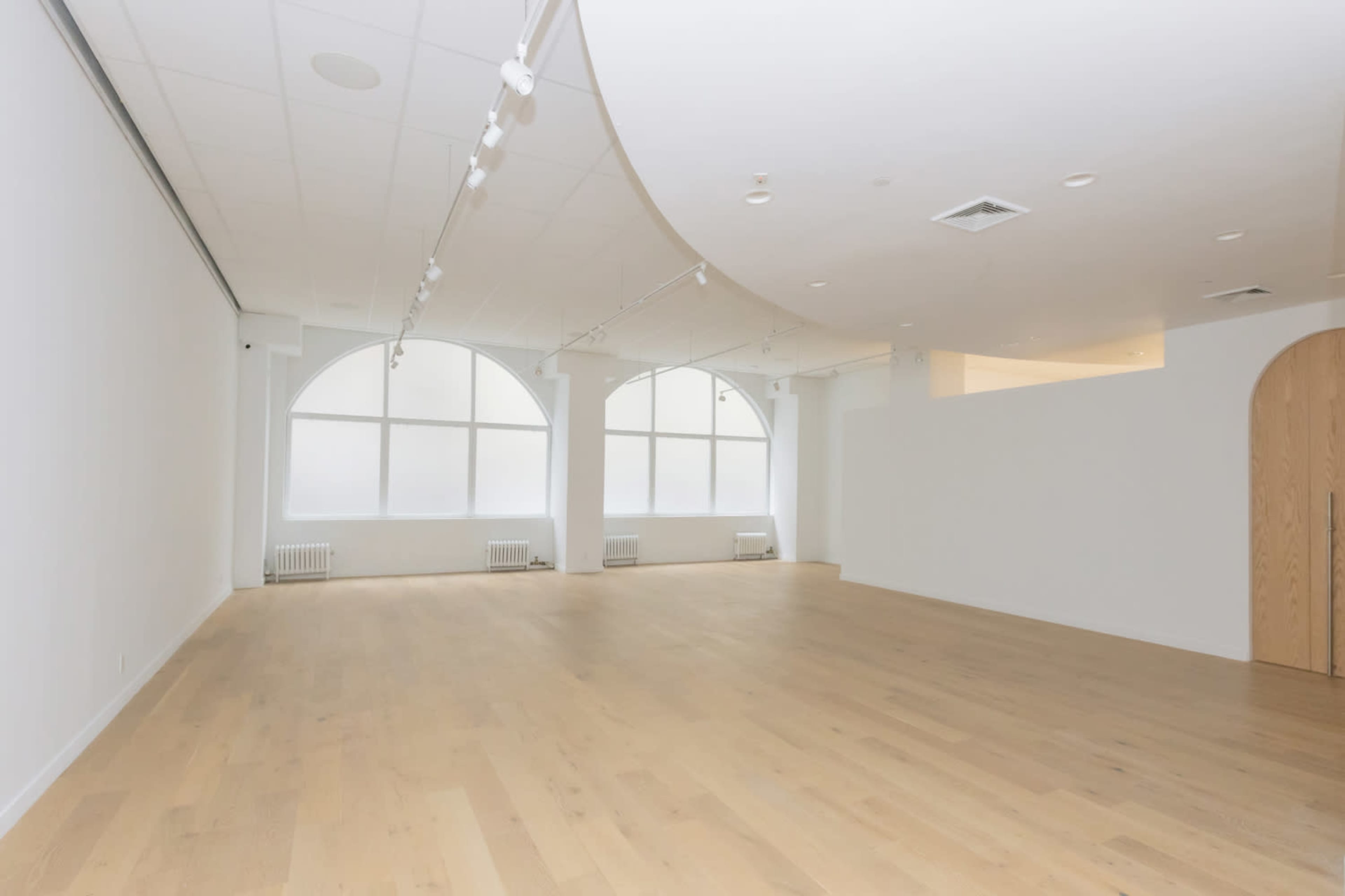 The image shows a spacious, empty room with large arched windows and a wooden floor.