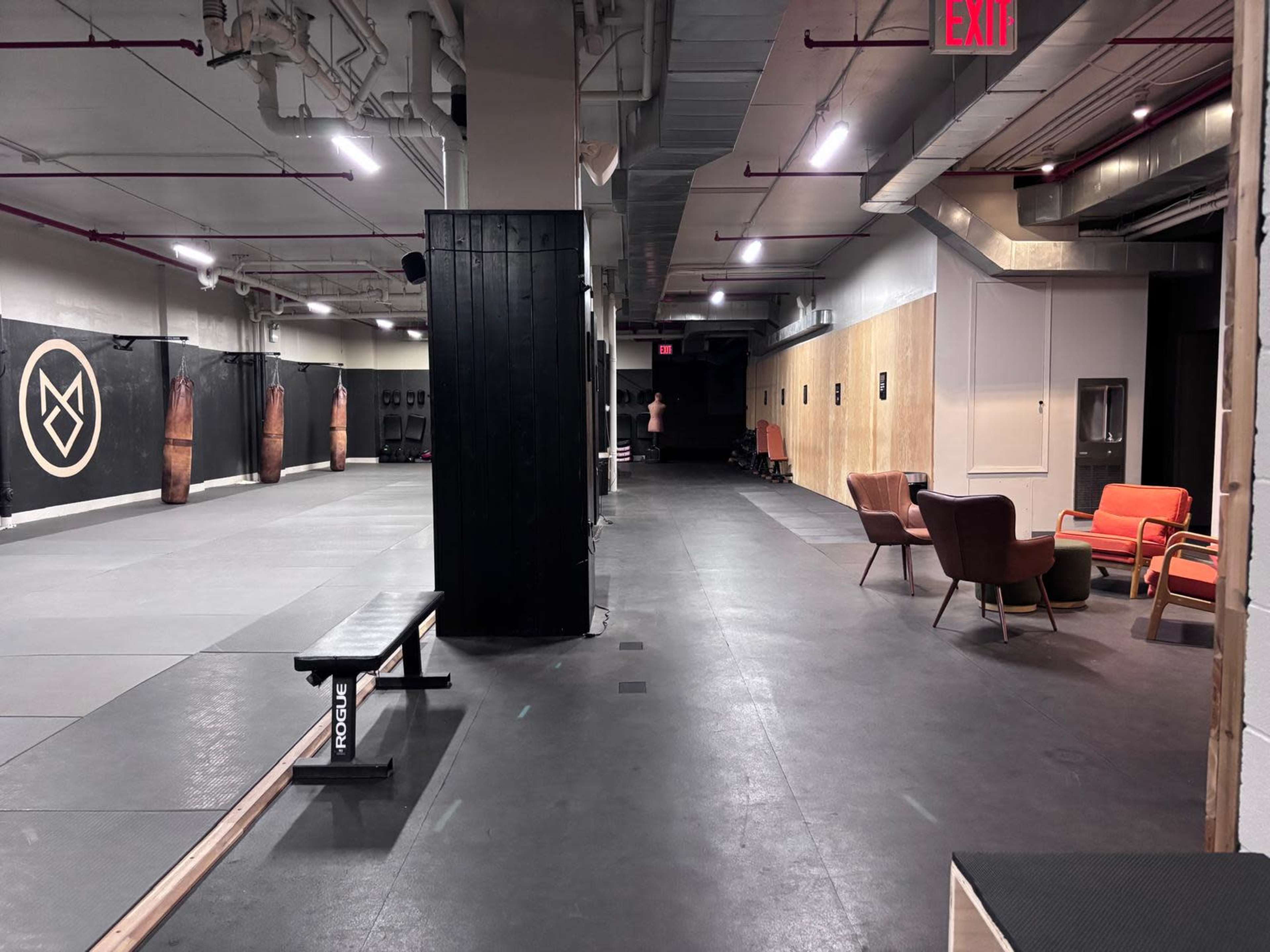 The image shows a spacious gym interior featuring workout mats, a seating area with colorful chairs, and boxing equipment along the walls.