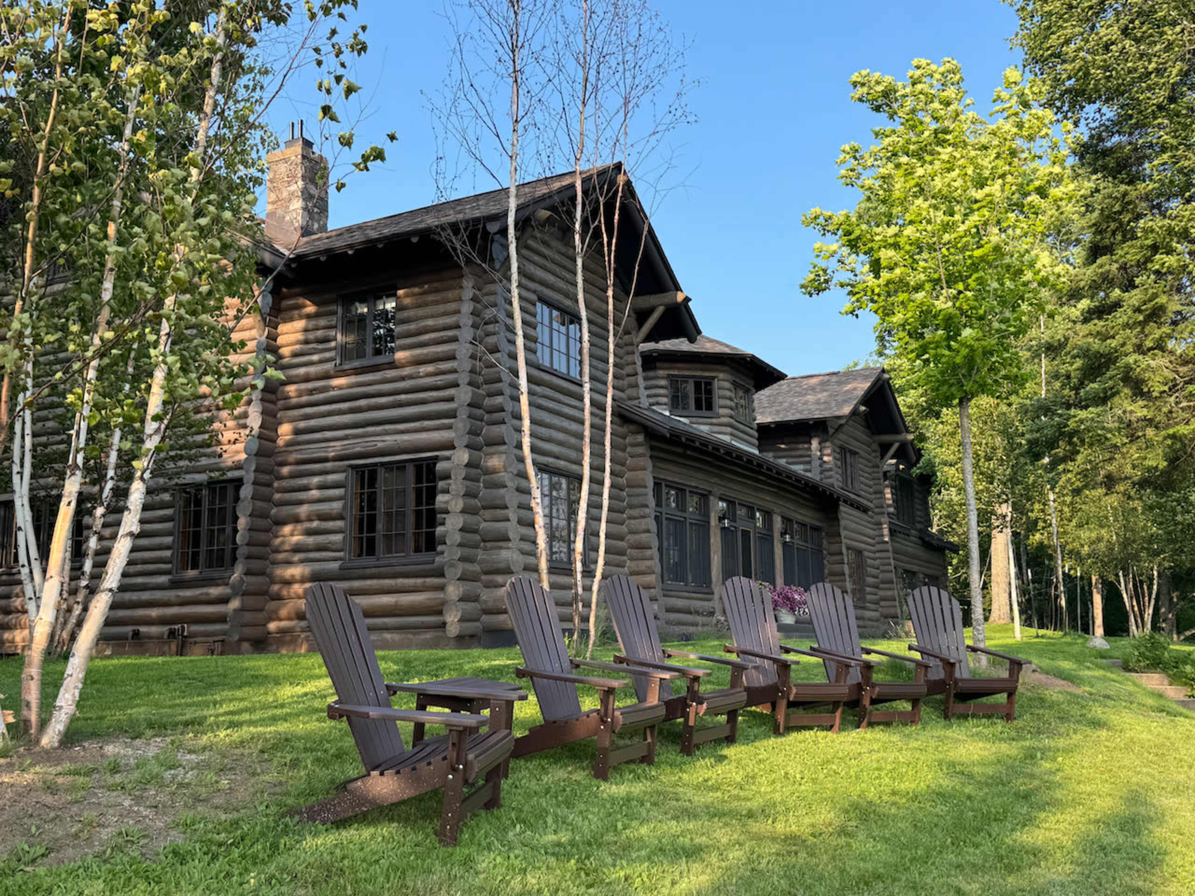 A log cabin with multiple windows is set in a green landscape, accompanied by a row of wooden adirondack chairs on the lawn.