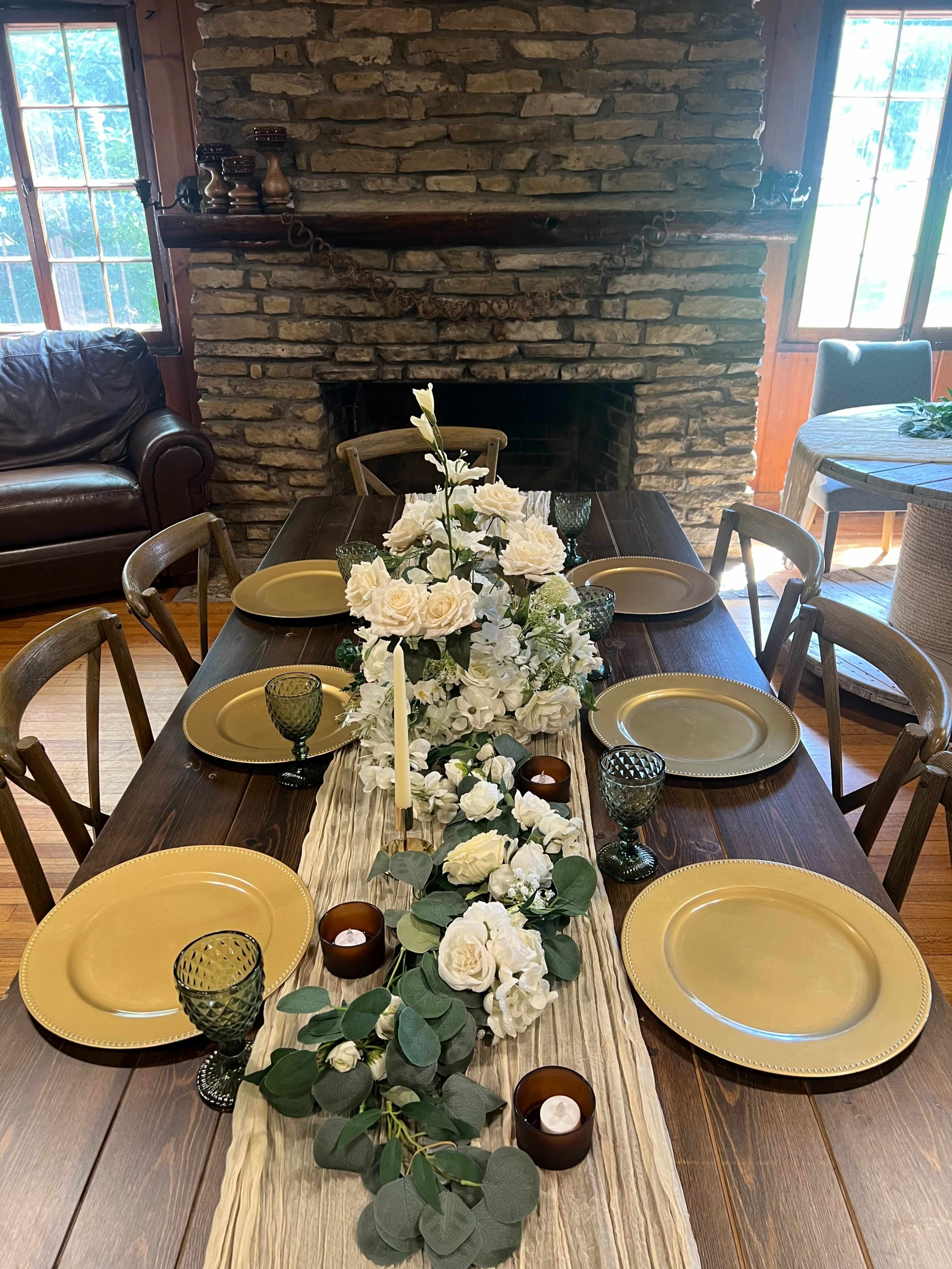 A wooden dining table is set with golden plates, green glassware, and a floral centerpiece, surrounded by chairs in a rustic room with a stone fireplace.