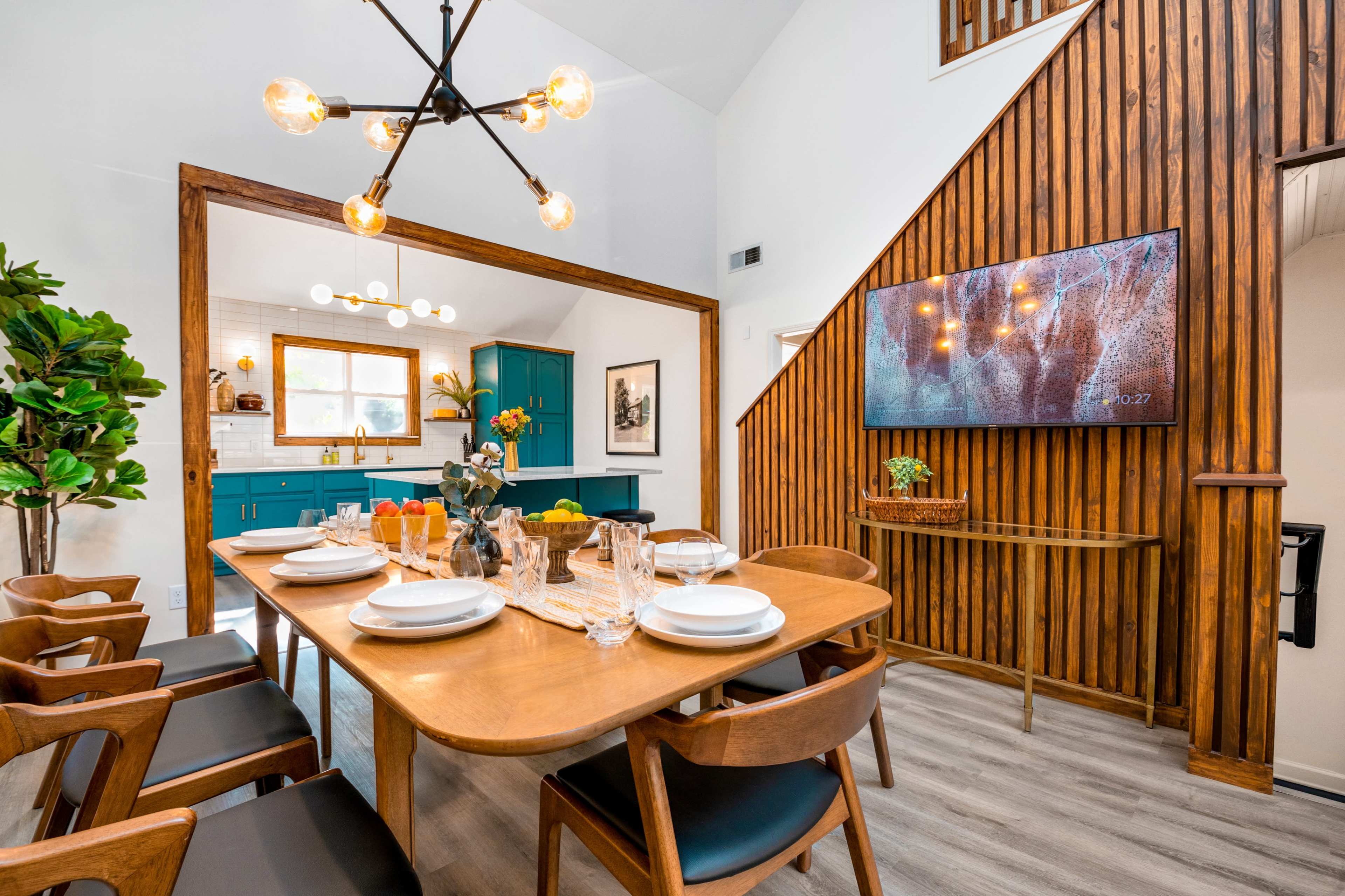 A dining area features a wooden table set for dinner, surrounded by chairs, with blue cabinets and a wall-mounted TV in the background.