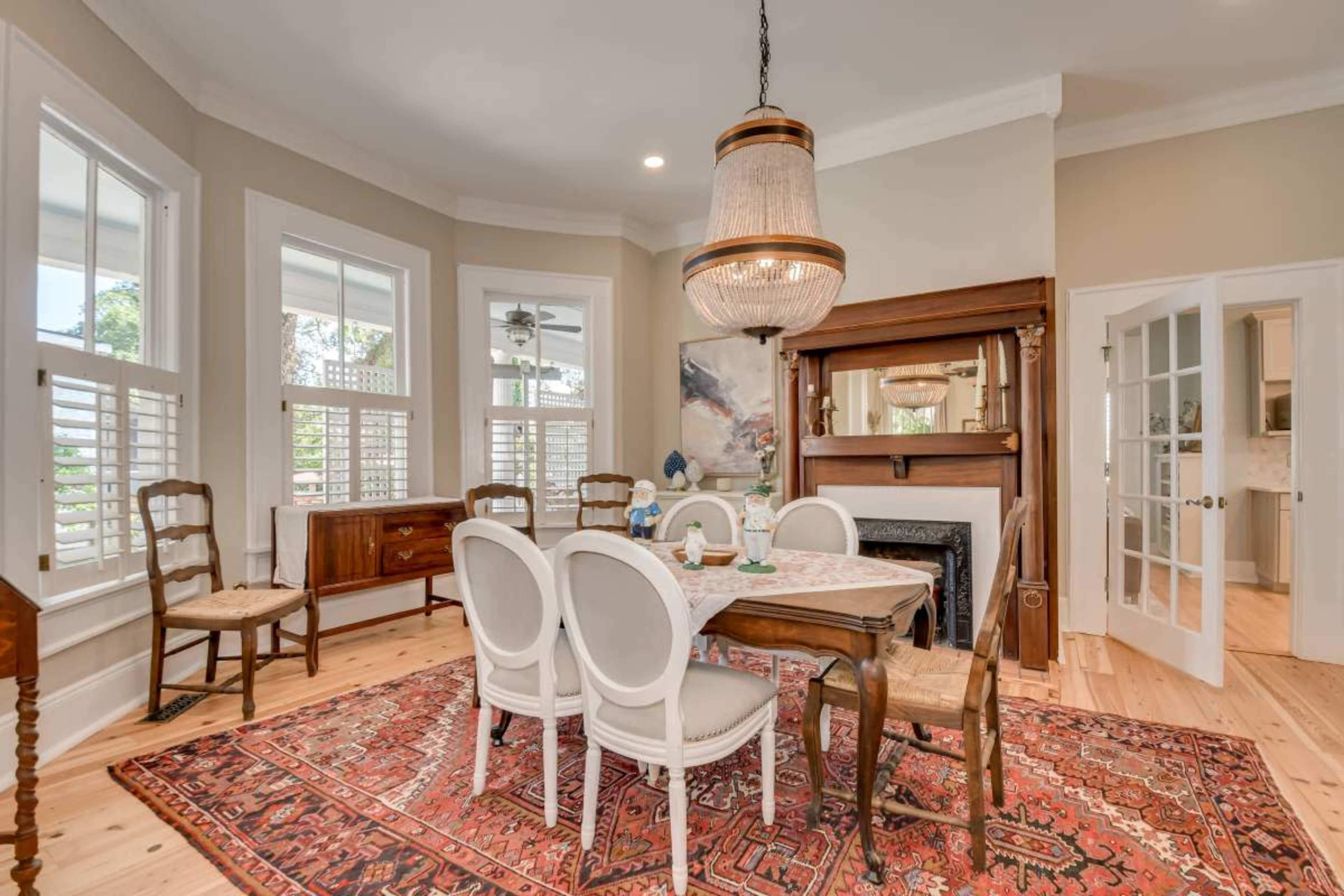 The image shows a dining room with a wooden table surrounded by chairs, a chandelier overhead, and large windows allowing natural light to enter.