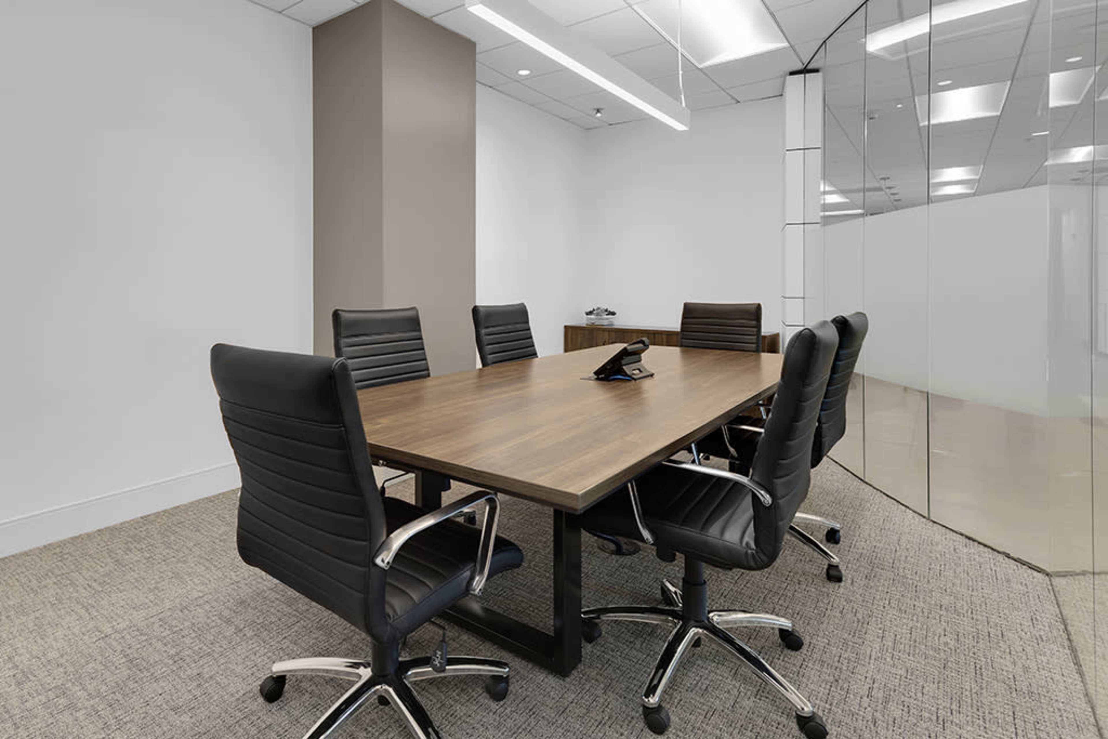 A conference room features a large wooden table surrounded by six ergonomic chairs, with a phone on the table and glass walls.