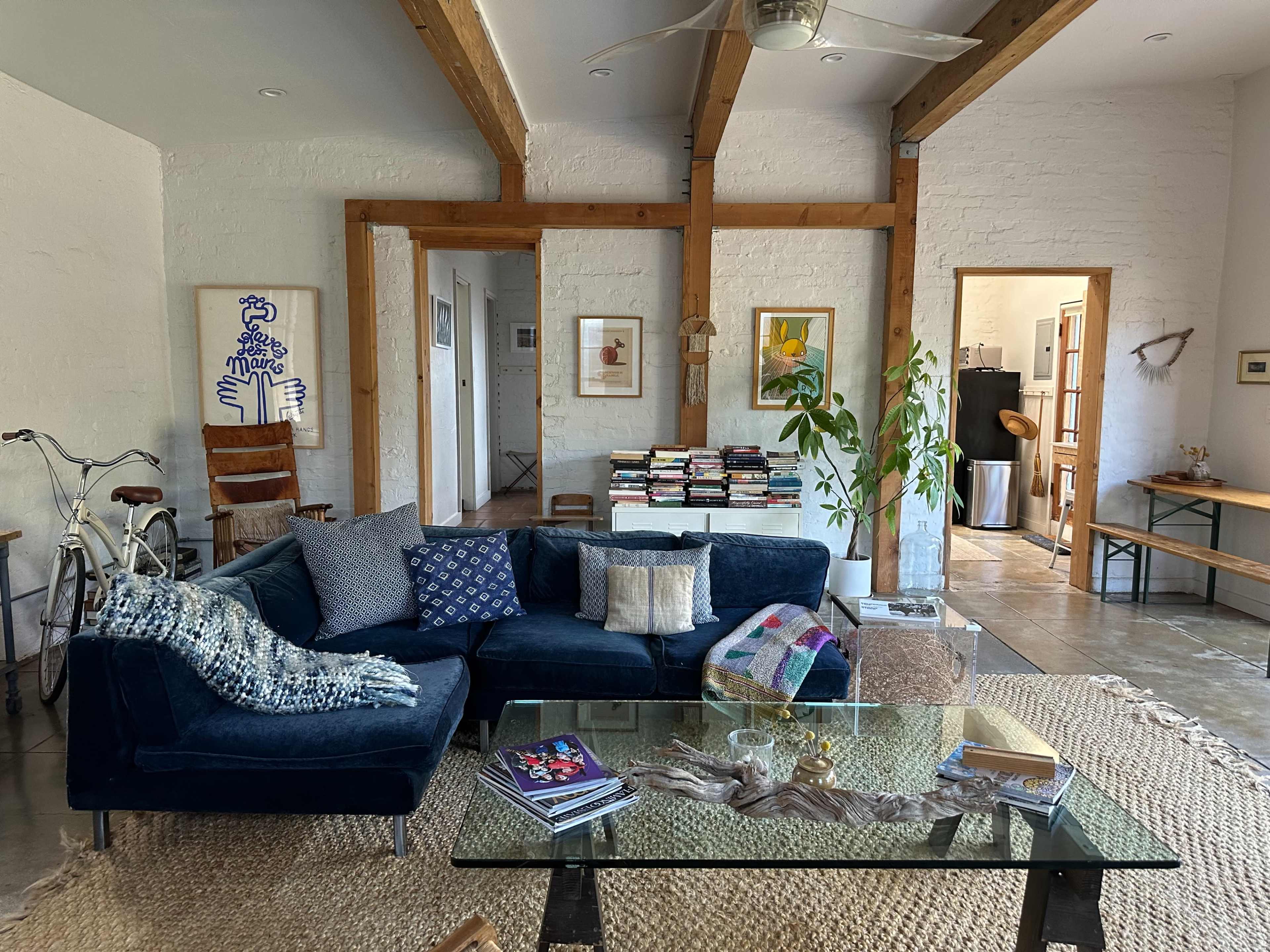A modern living room with a blue sofa, a glass coffee table, a bookshelf, and decorative plants, featuring exposed wooden beams and light-colored walls.