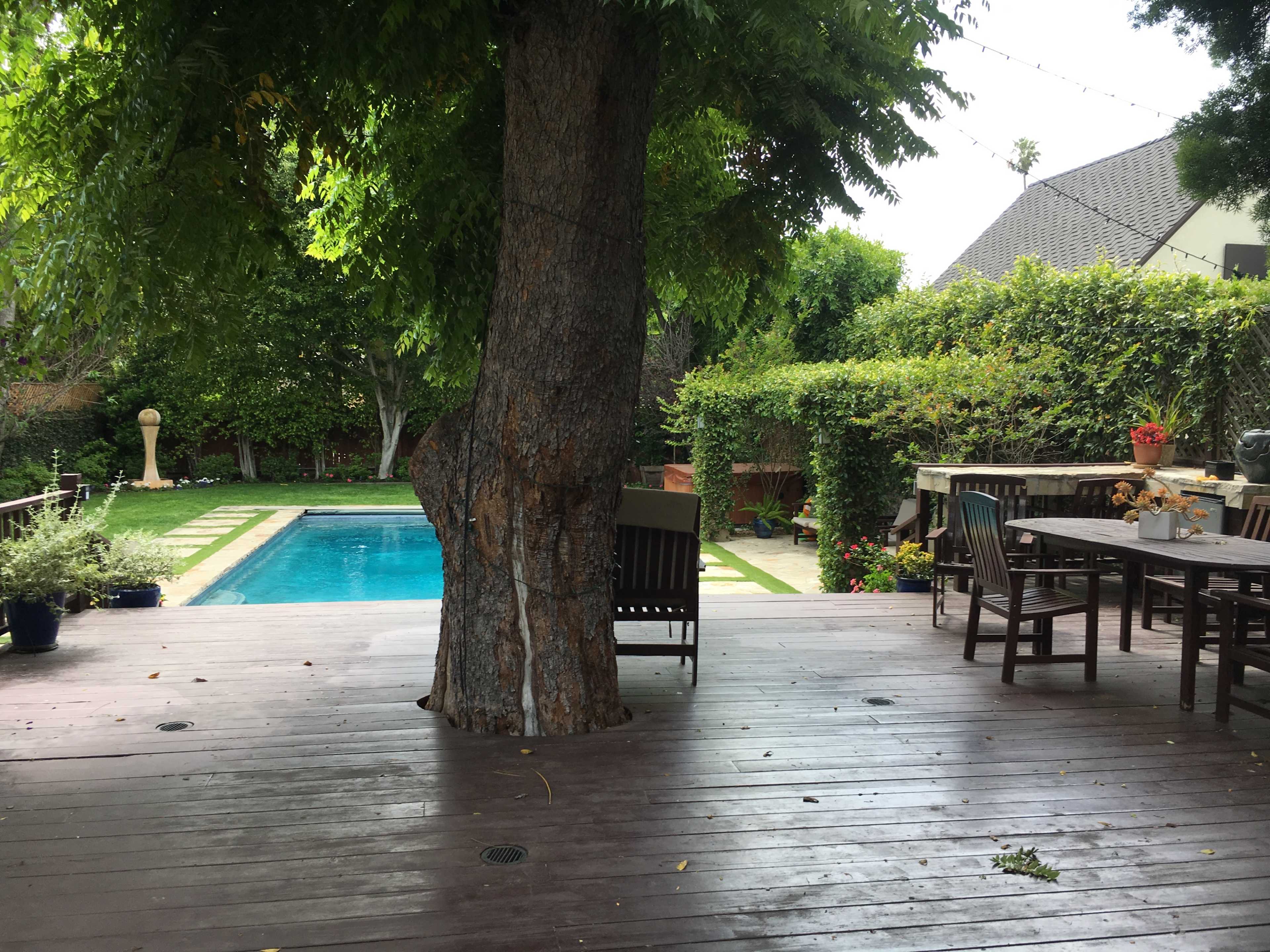 A wooden deck with several chairs surrounds a swimming pool, set against a backdrop of lush greenery.