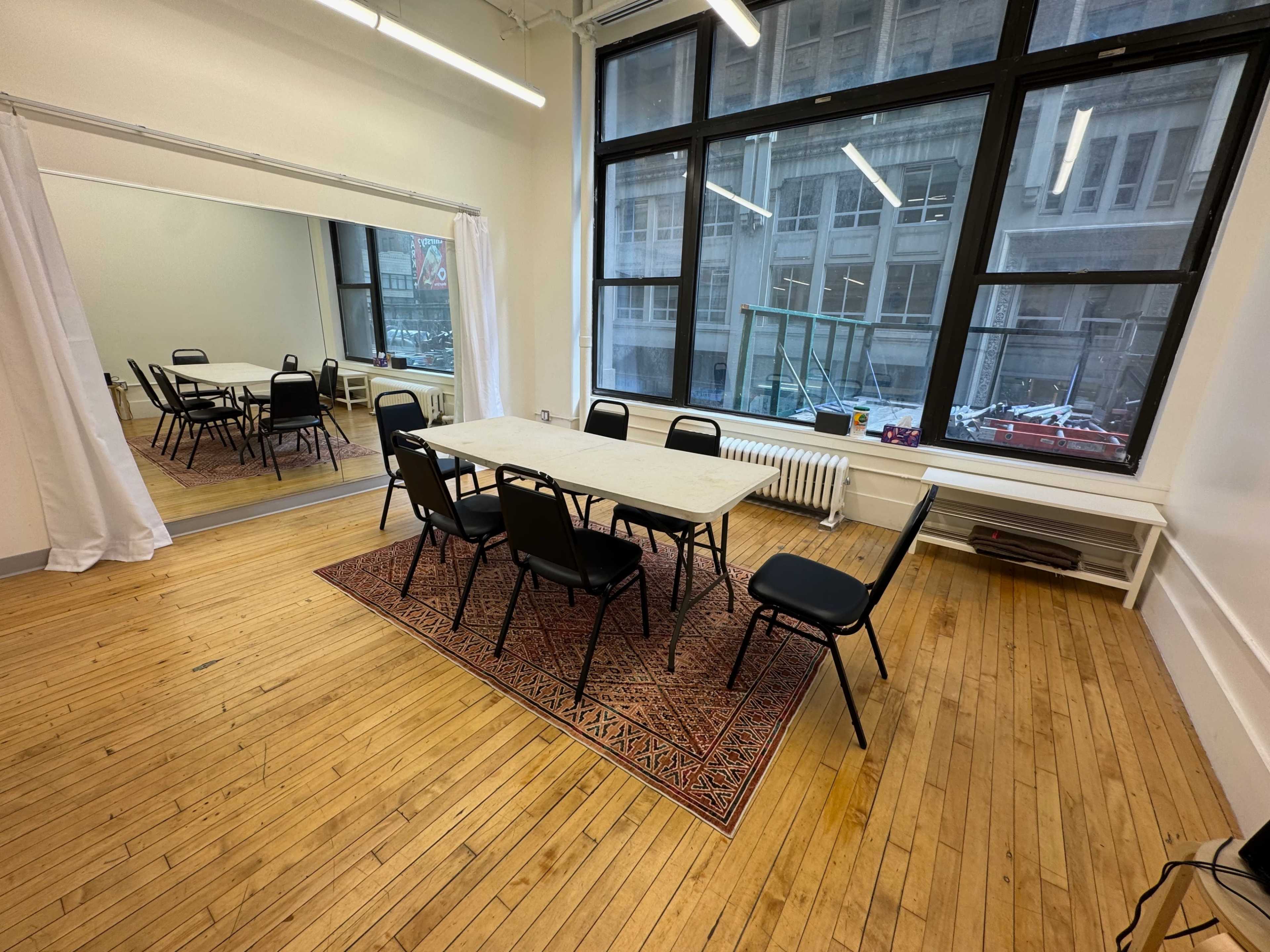 The image shows a meeting room with a rectangular table surrounded by black chairs, a large mirror on one wall, and large windows letting in natural light.