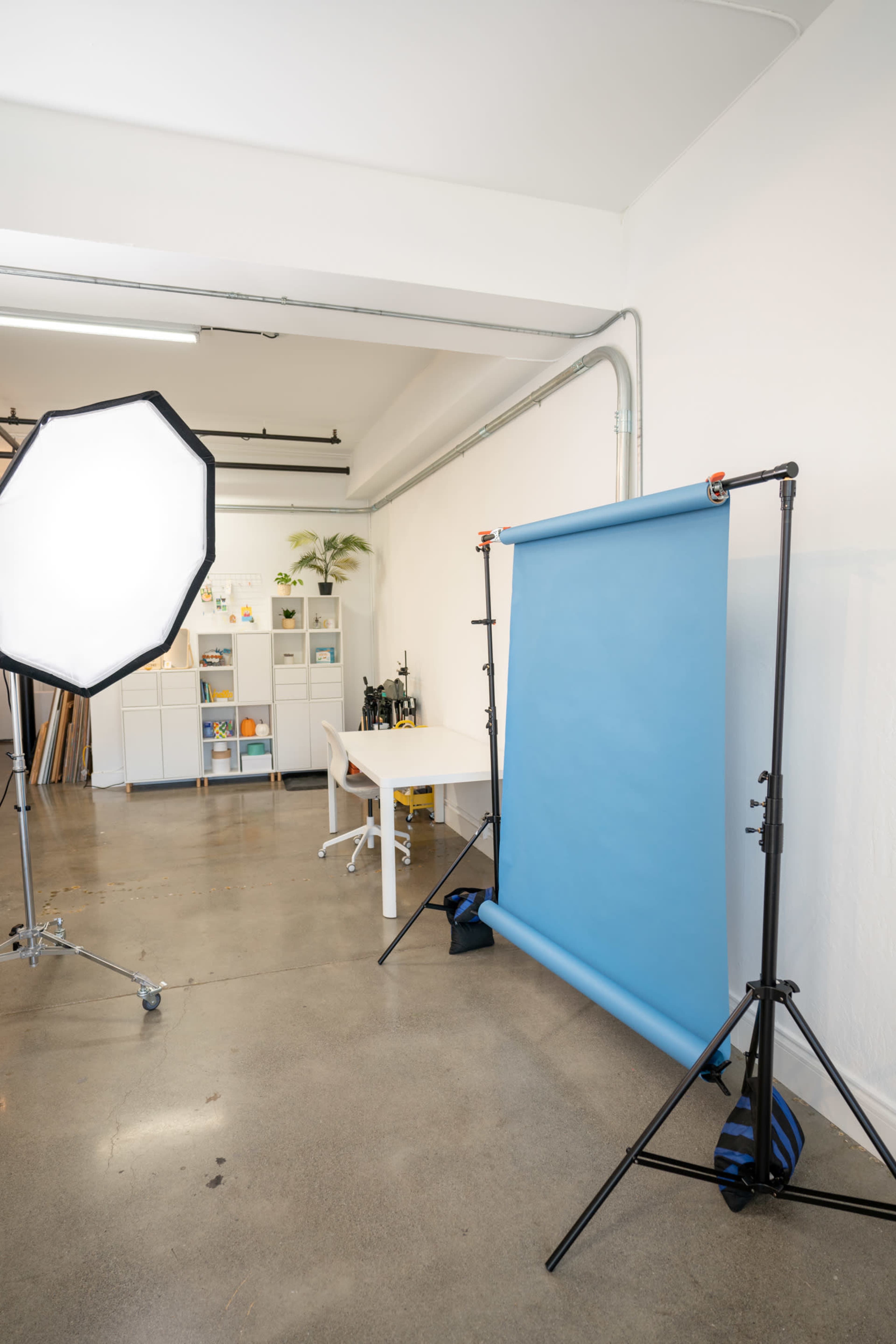 A photography studio features a blue backdrop, a large light source, and a white desk with shelves in the background.