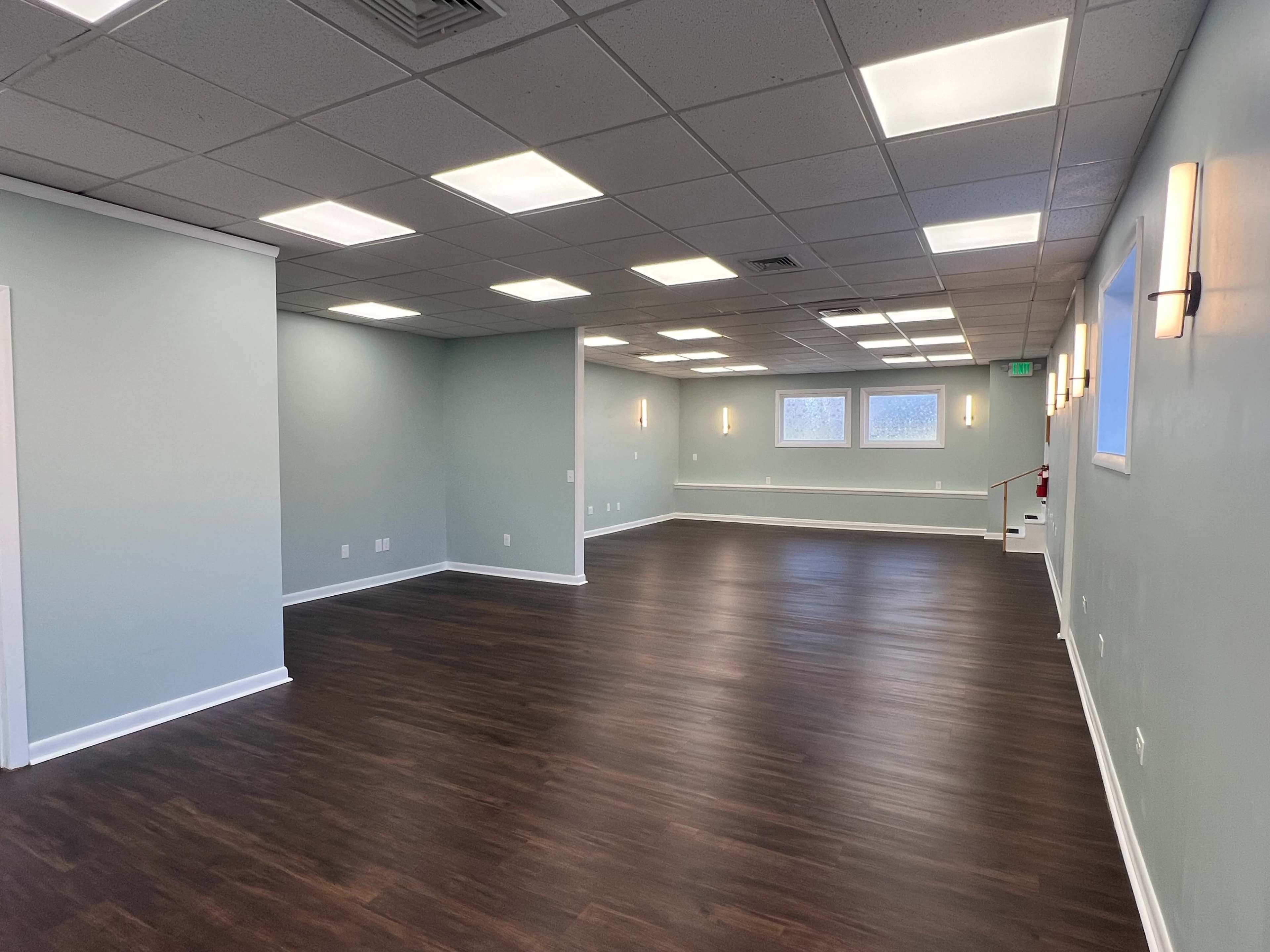The image shows a spacious, empty room with gray walls and wooden flooring, illuminated by overhead lights and wall sconces.