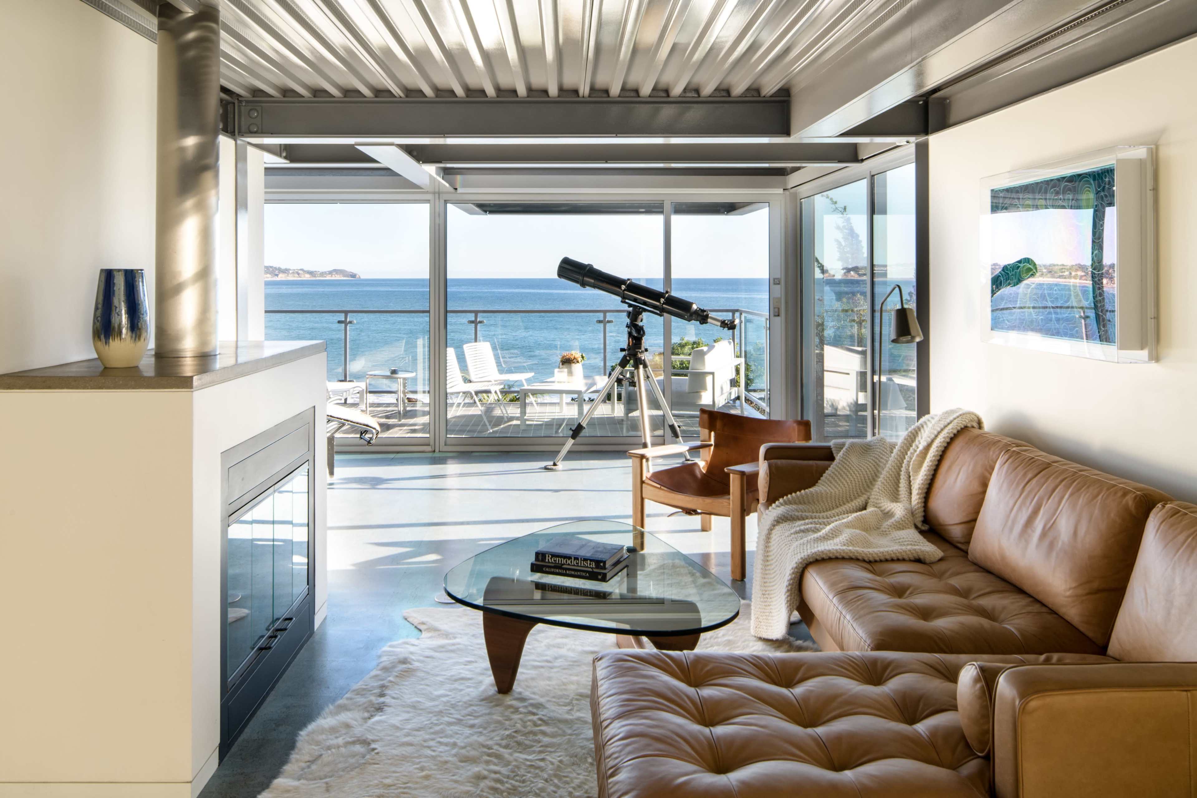 The living room features a leather sofa, a glass coffee table, and a telescope, all overlooking a scenic ocean view through large glass windows.