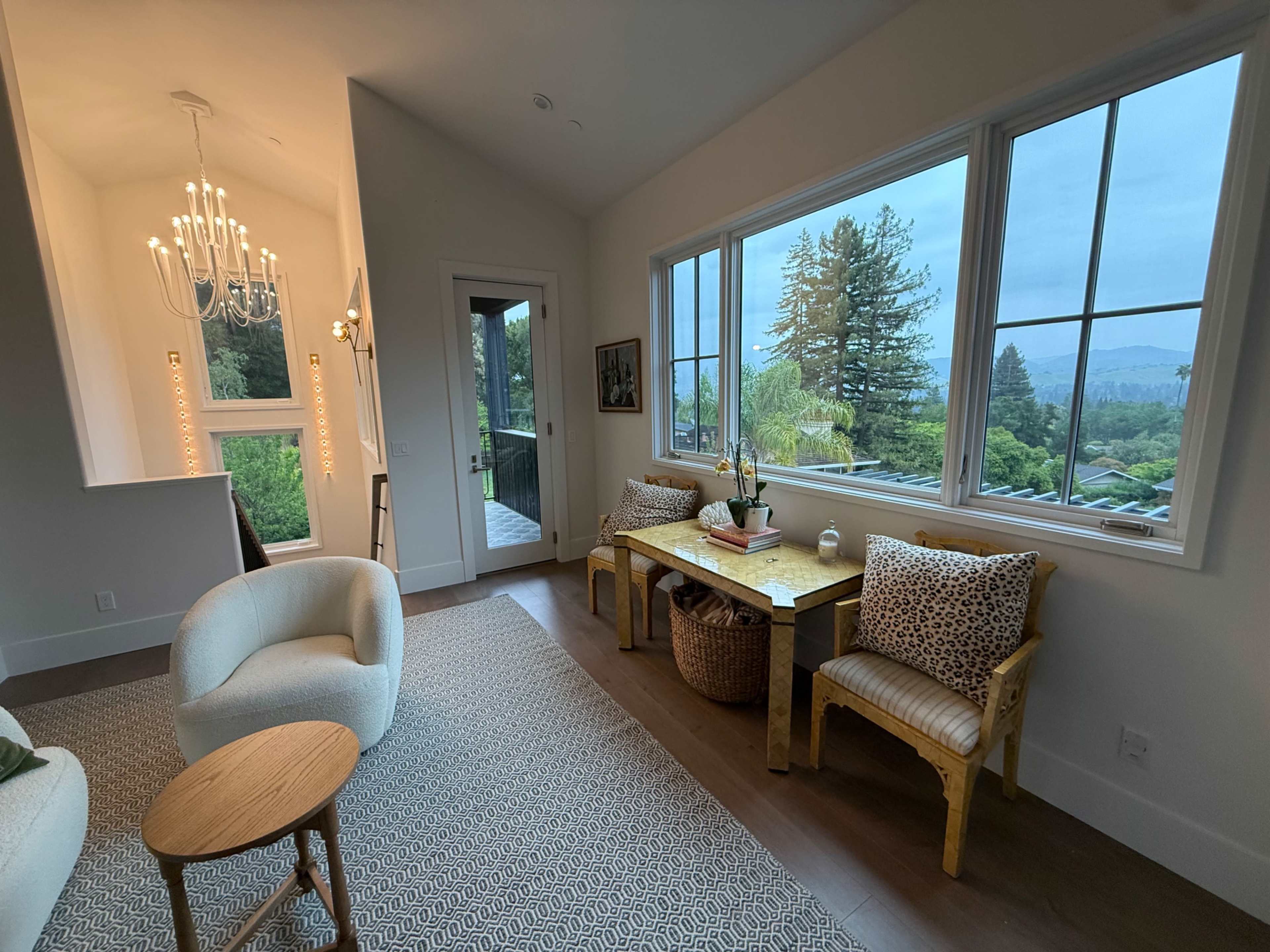 A bright living space features a small dining area with two patterned chairs, a wooden table, and large windows showcasing a view of trees and mountains.