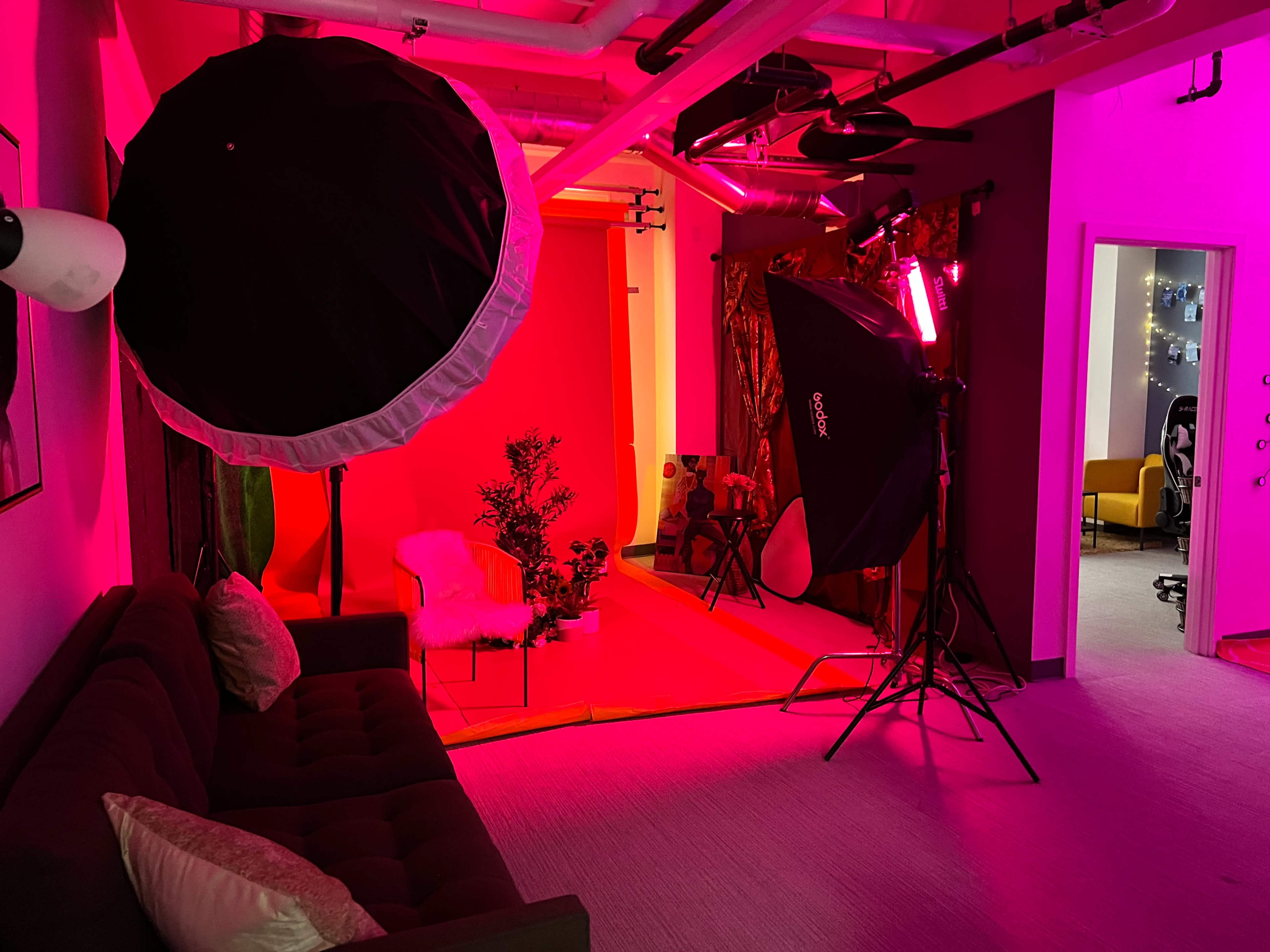The image shows a photography studio with a red backdrop, lighting equipment, and a brown couch in a brightly colored space.