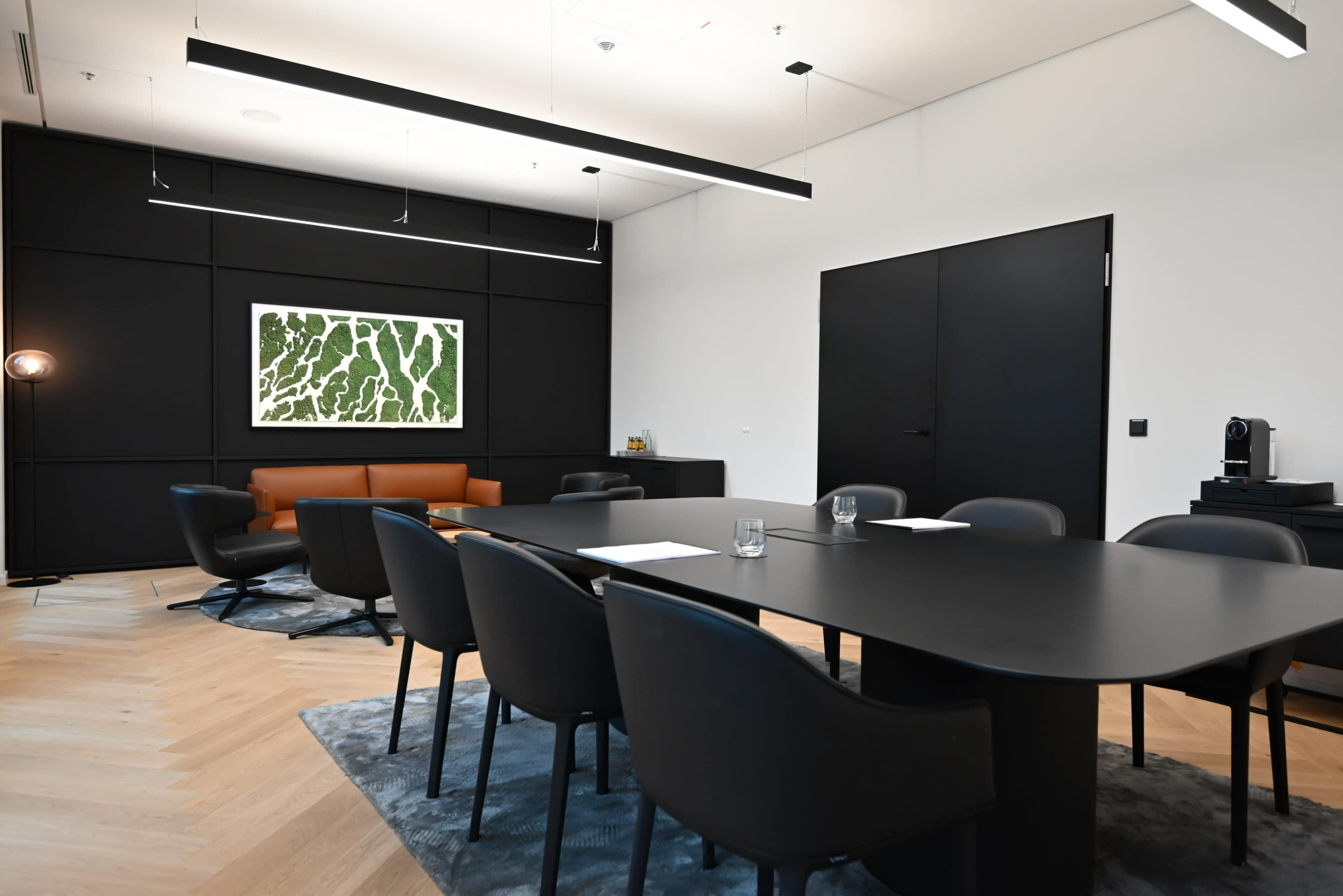 A modern conference room features a large black table surrounded by ergonomic chairs, a brown leather sofa against the wall, and a framed artwork of green patterns.