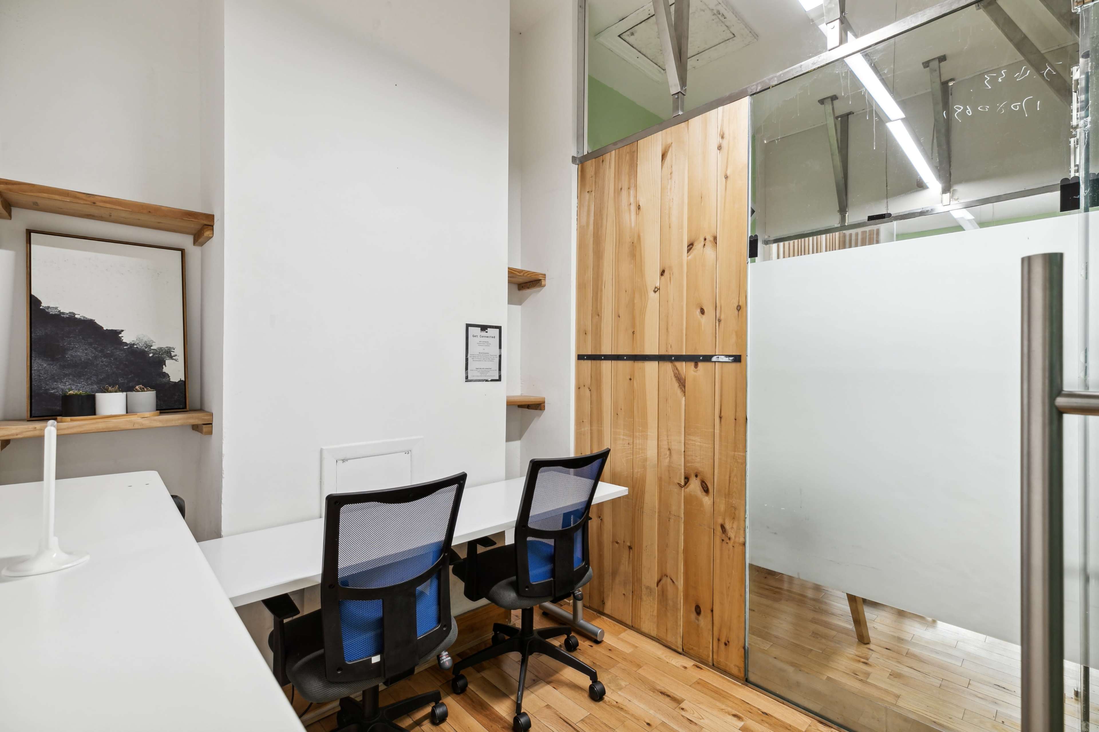 A small office space with two chairs at a white desk, a wooden board separating a glass-walled meeting room, and shelves mounted on the wall.