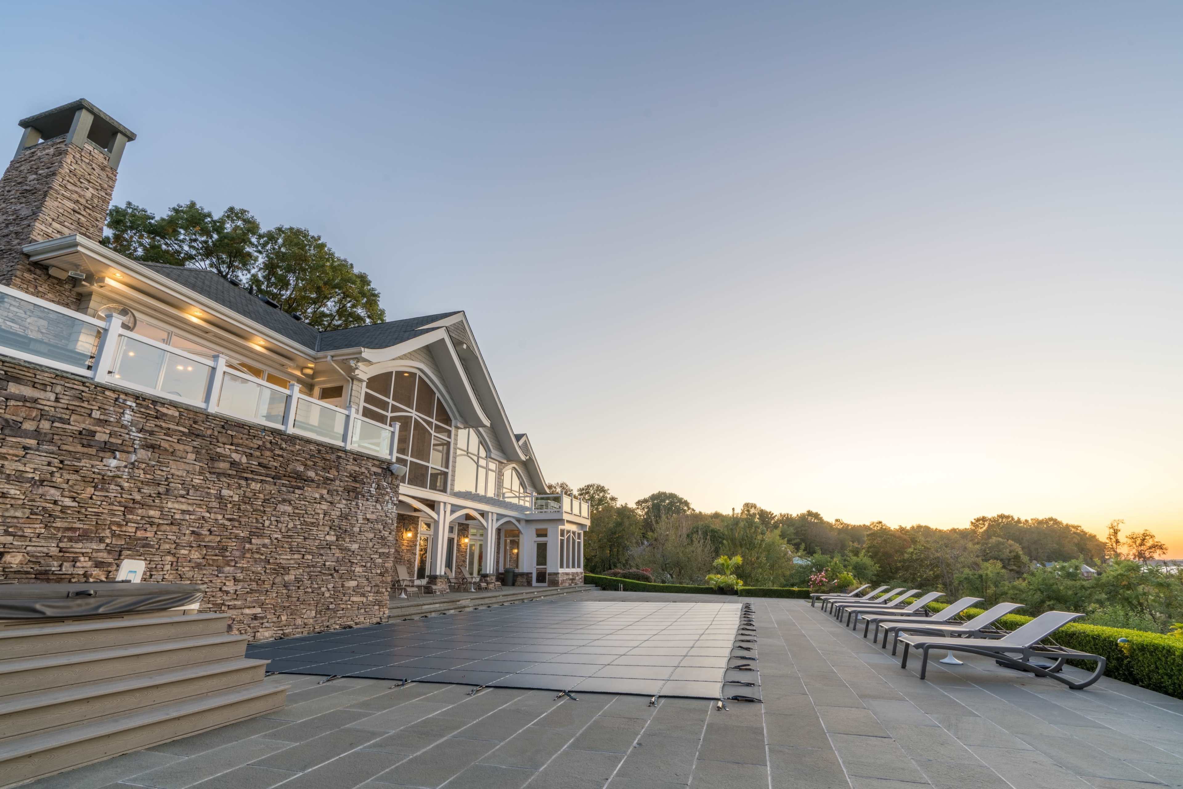 A large stone house with multiple windows overlooks a covered swimming pool and several lounge chairs on a patio.