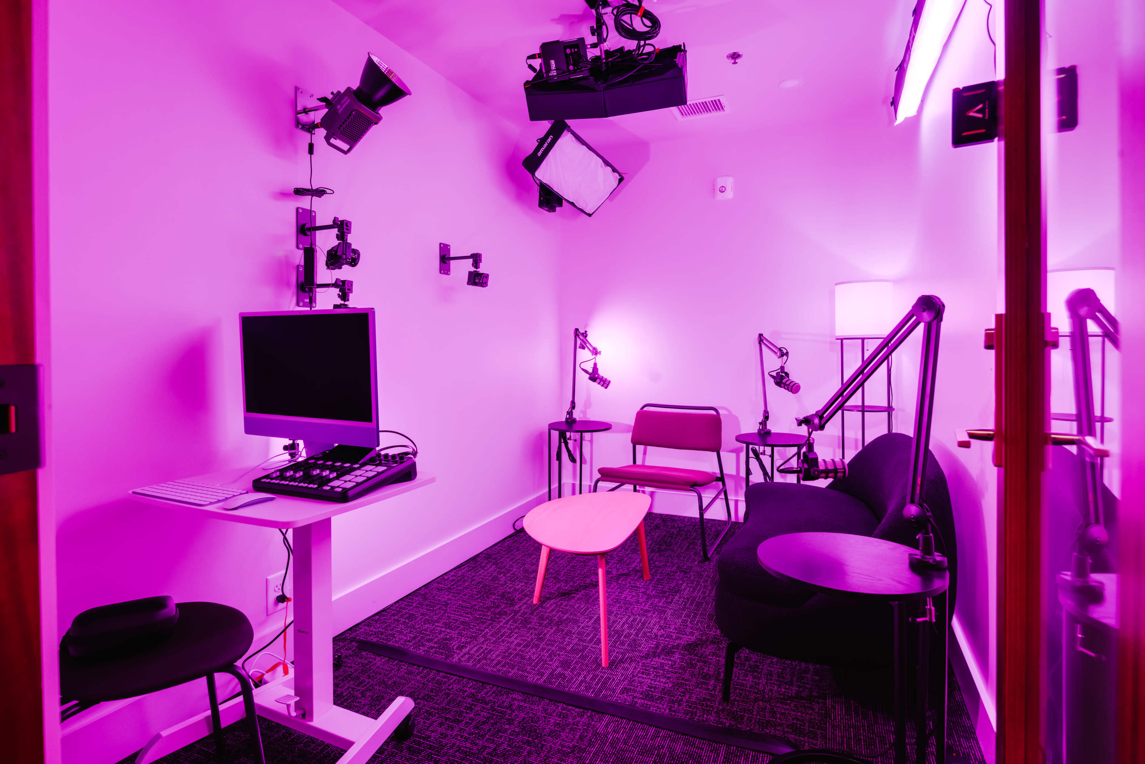 The image shows a small studio room illuminated with pink lighting, featuring a computer desk, chairs, and various lighting equipment mounted on the walls and ceiling.