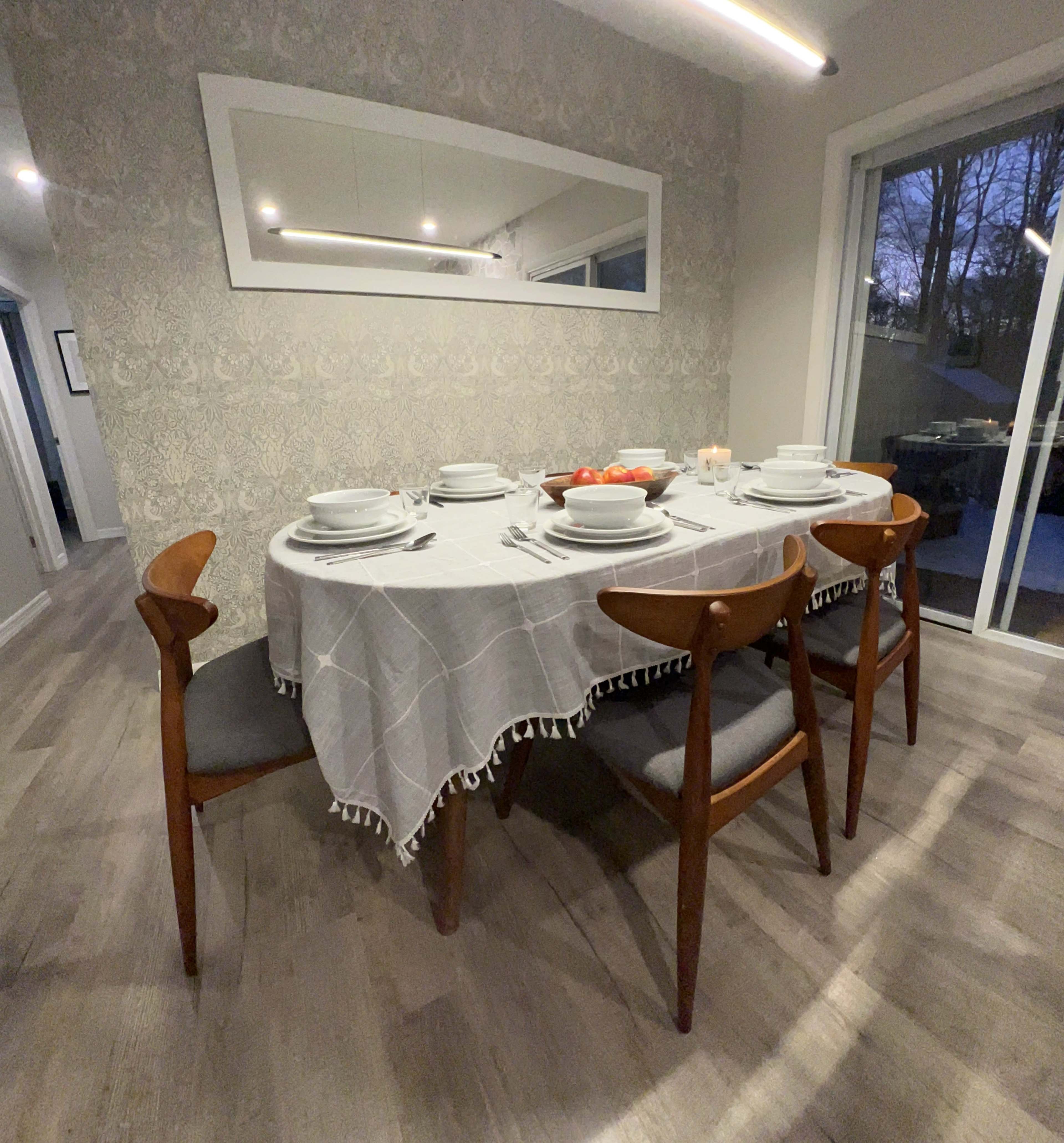 A dining table set with white plates and a light gray tablecloth is arranged with wooden chairs in a well-lit room.