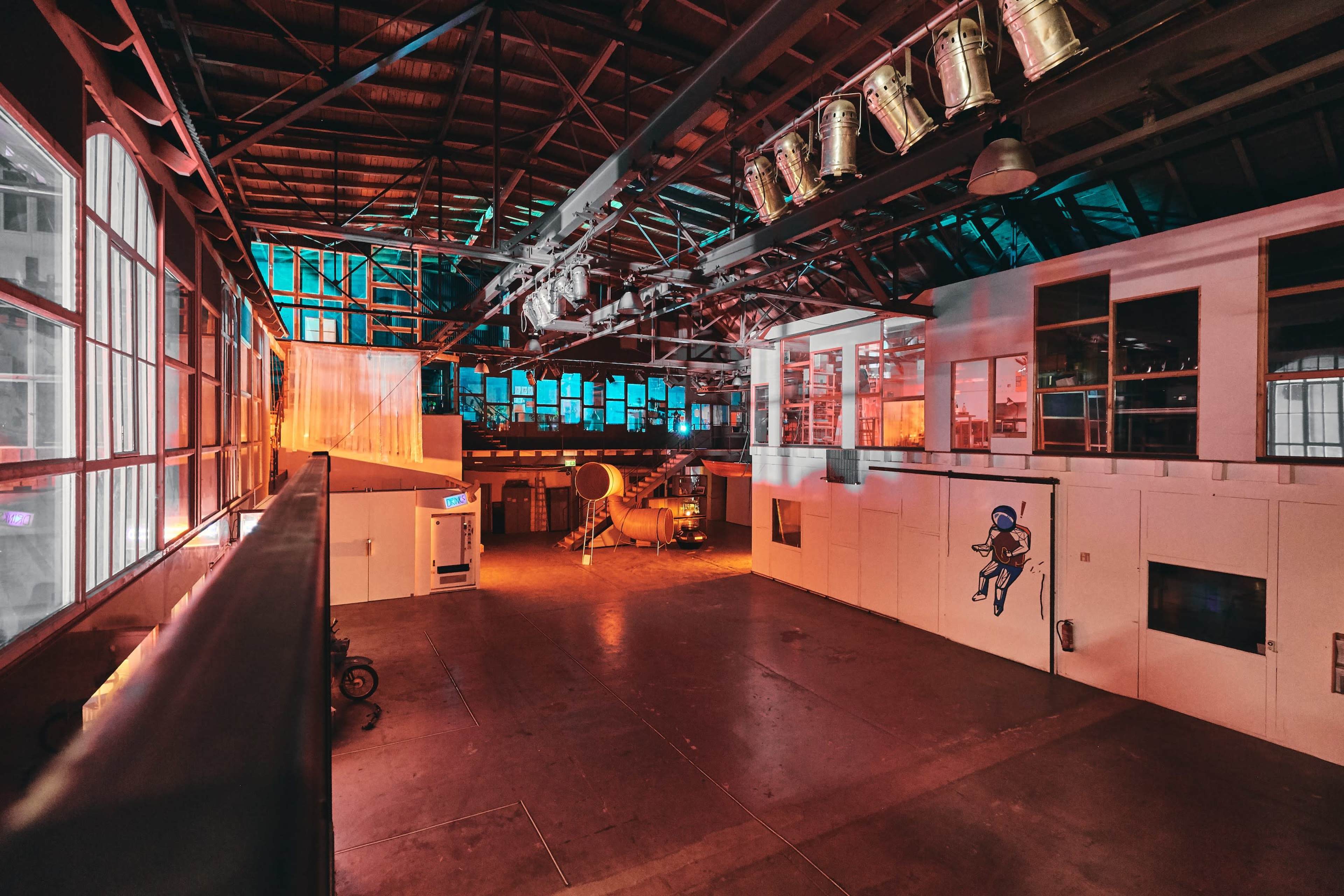 A spacious industrial hall with large windows, orange and blue lighting, and various equipment along the walls.