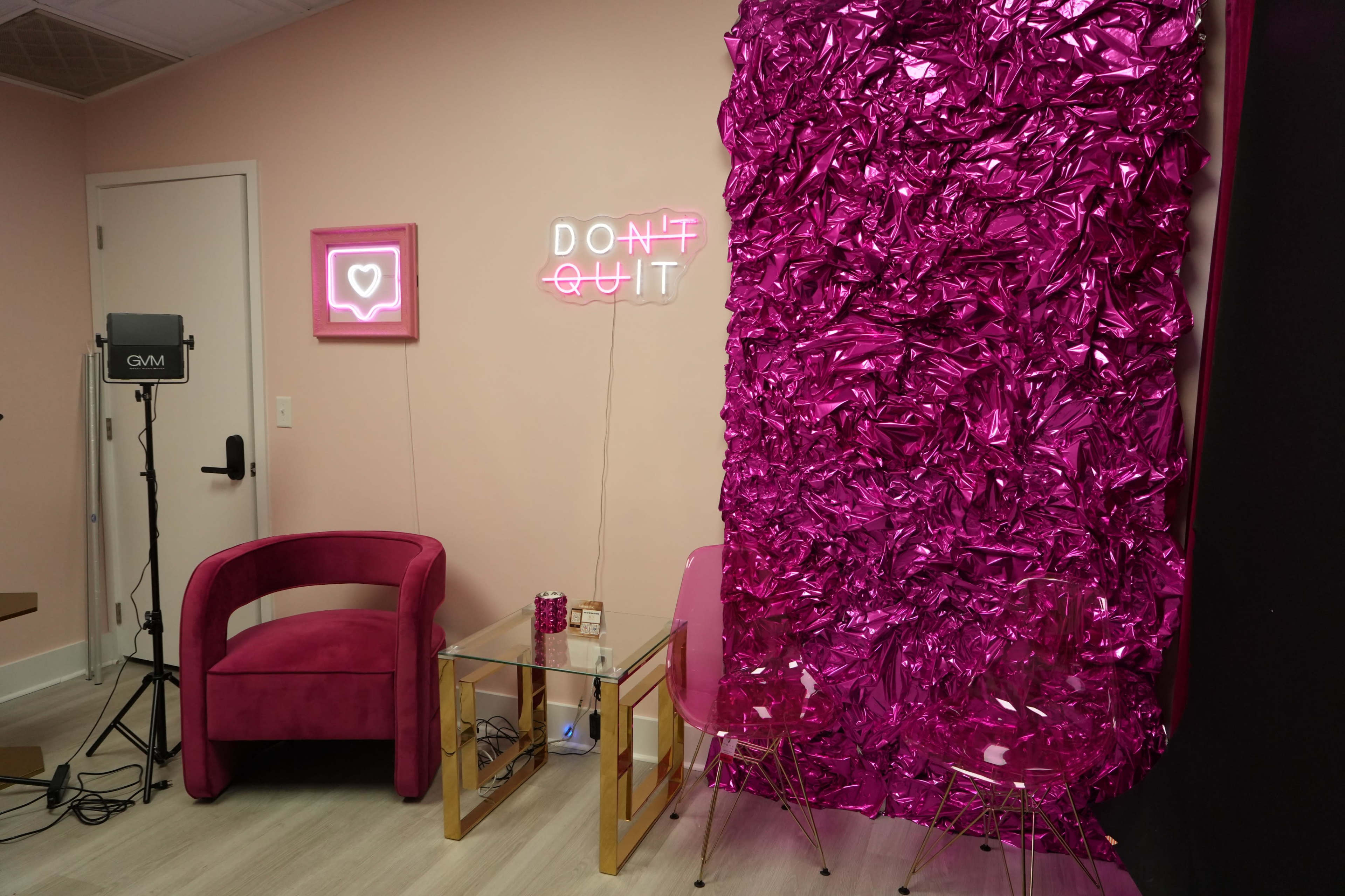 A pink-themed room features a plush chair, a glass table, and a wall decorated with crumpled pink material, accented by neon signs.