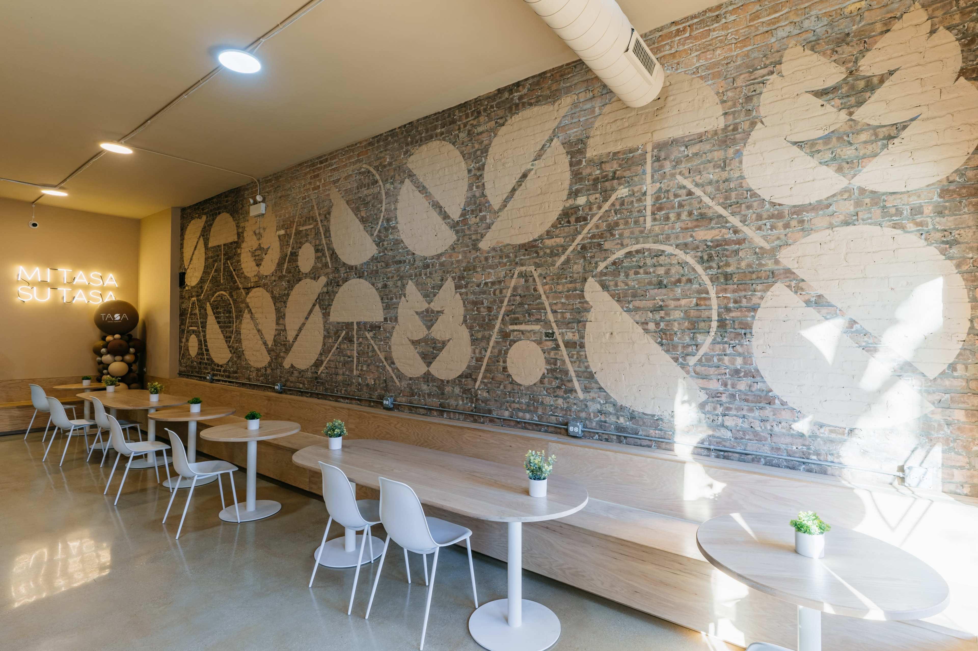 The image shows a modern interior with wooden tables and white chairs, set against a brick wall featuring a large abstract mural and illuminated signage.